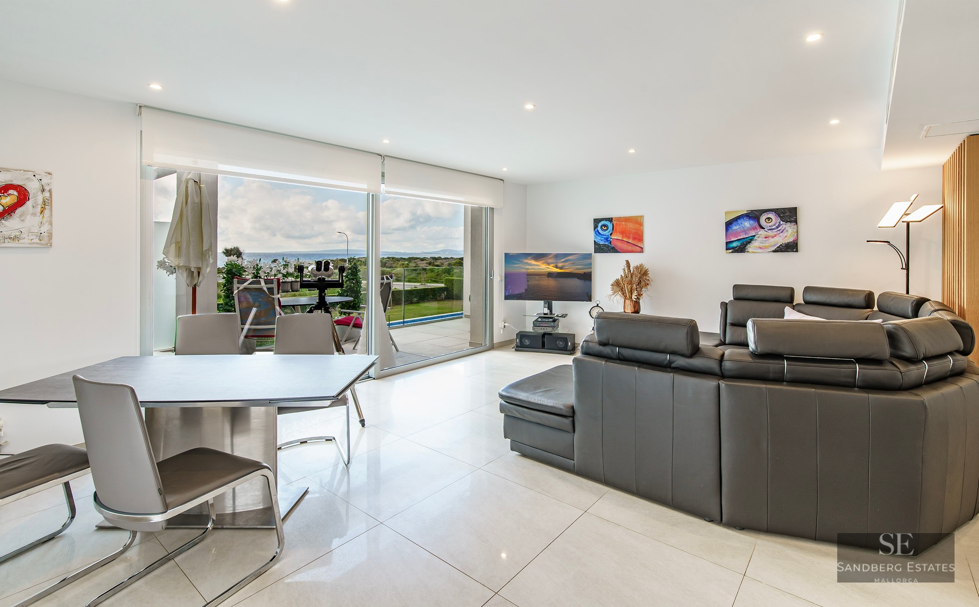 Modern living room with dark grey leather sofa, dining area, and large glass doors opening to a sea view.