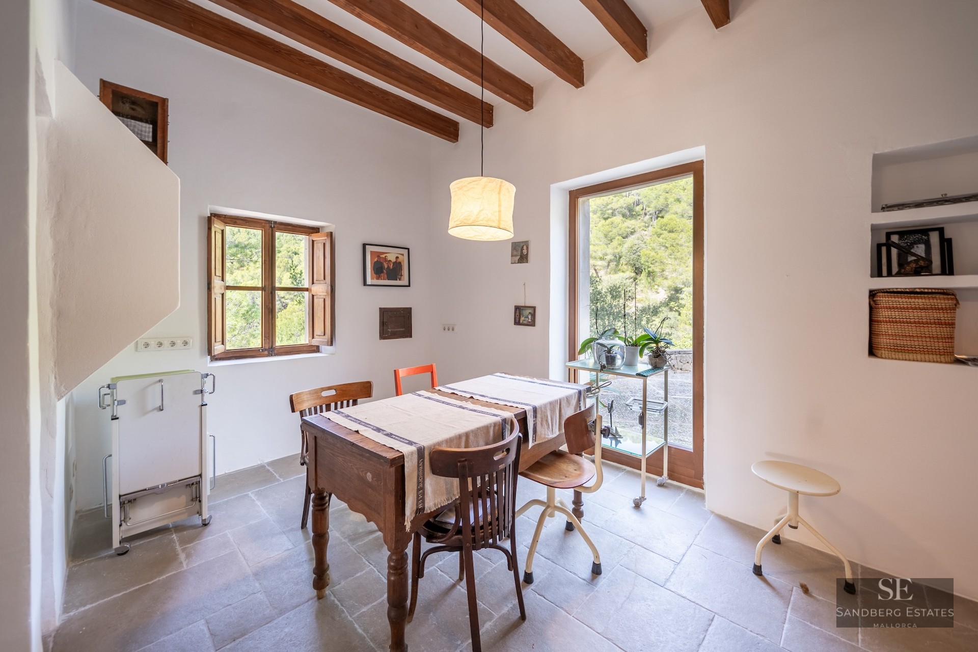 Bright dining room featuring a wooden table, mismatched chairs, exposed ceiling beams, and a glass door overlooking a forest.