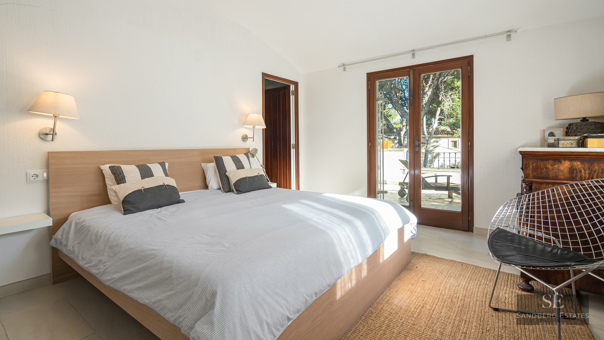 Bright bedroom with a wooden bed, white walls, and glass doors opening to a sunlit terrace.
