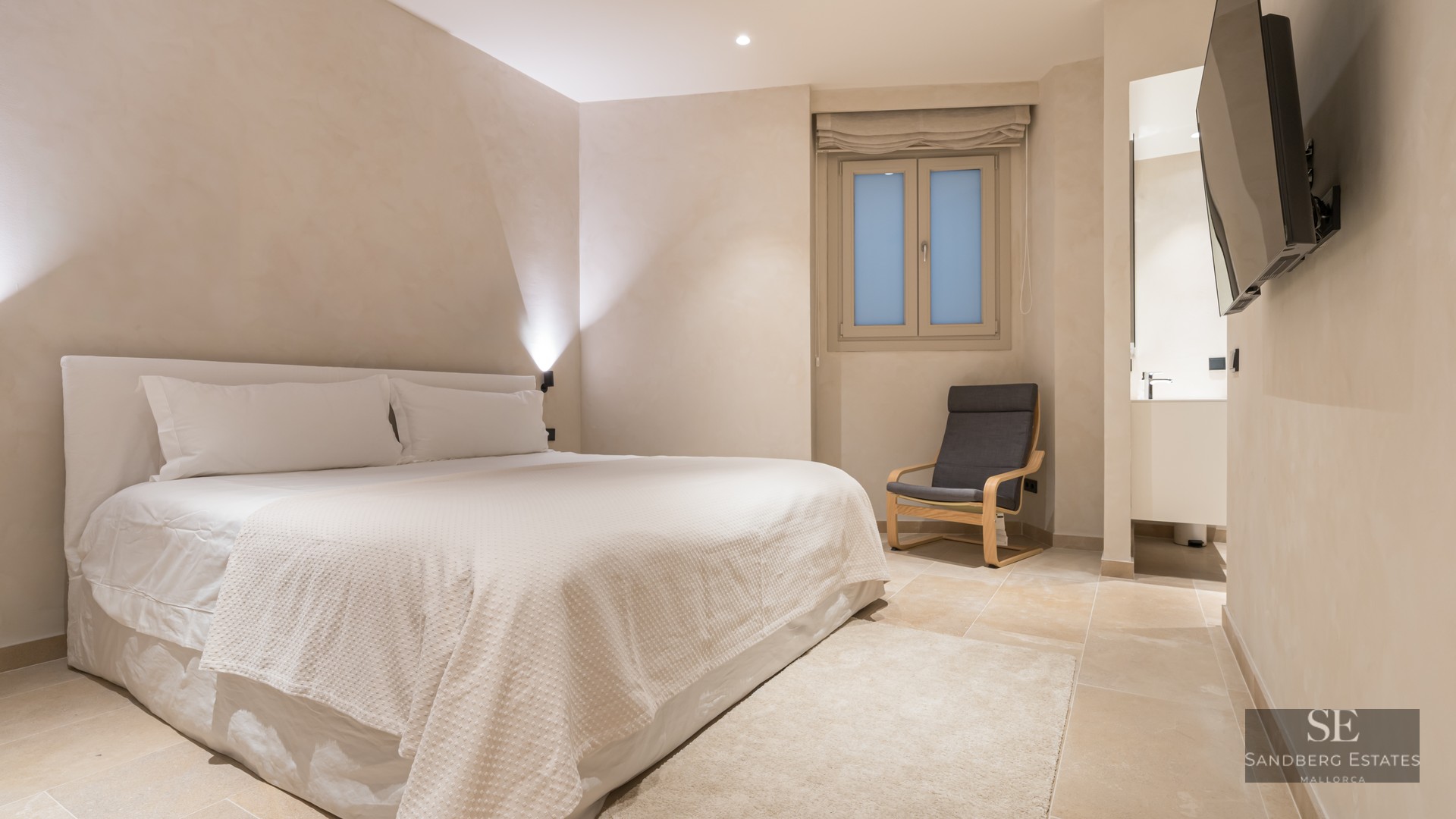 A serene bedroom with a large white bed, a grey designer chair, stone flooring, and soft beige walls.