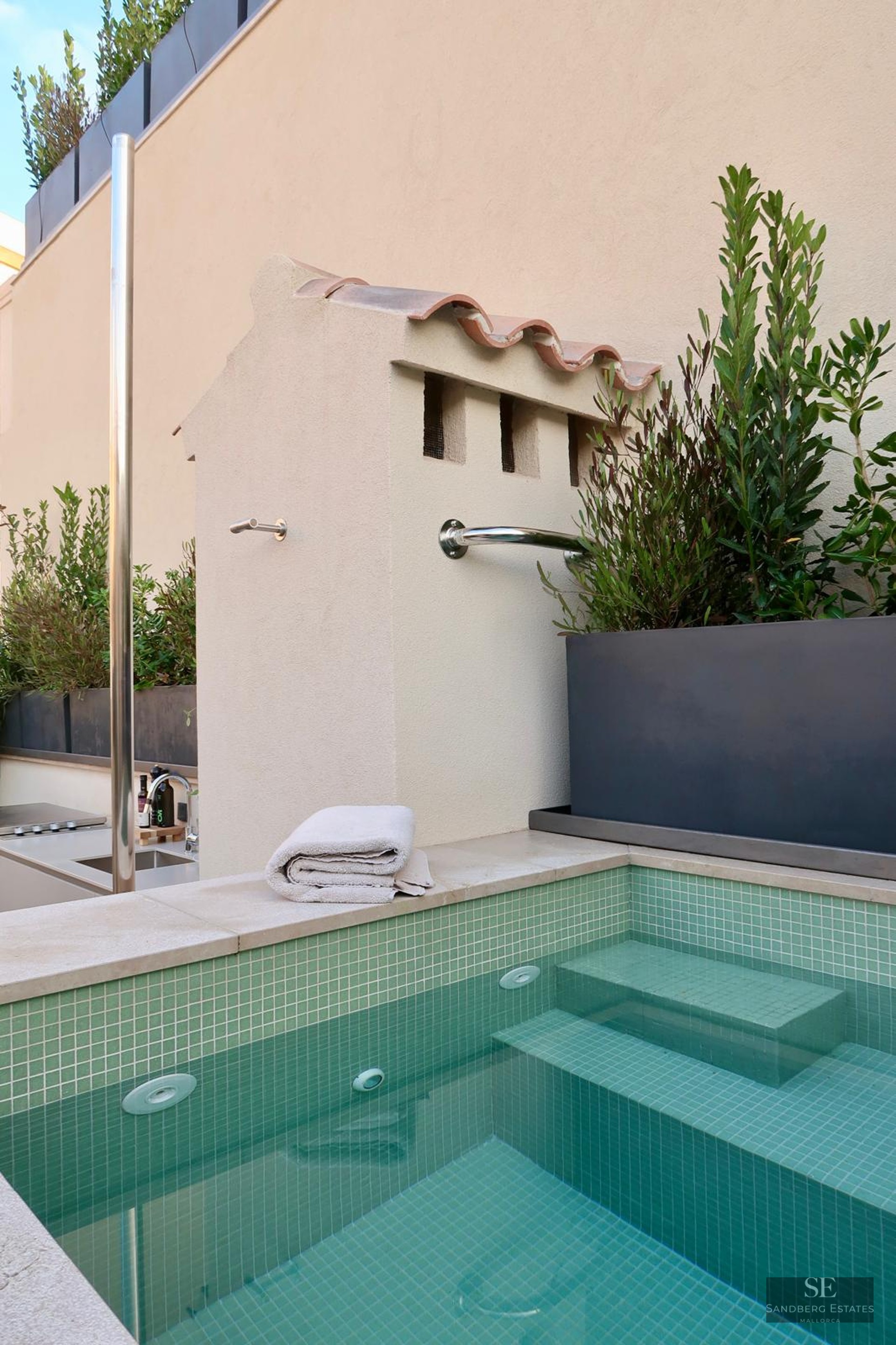Green mosaic tile pool with submerged steps and a stainless steel shower next to a light-colored wall.