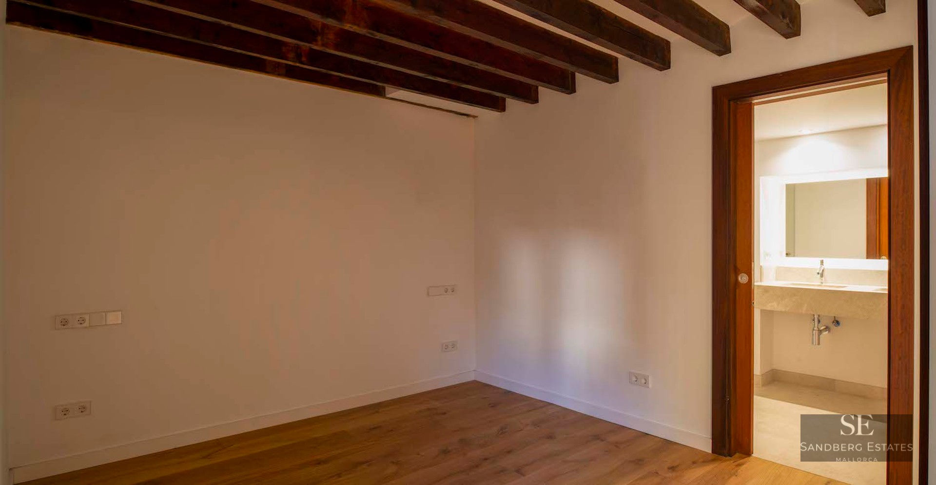 Empty bedroom with dark wood ceiling beams, hardwood floors, and a doorway leading to a modern bathroom with a stone vanity.