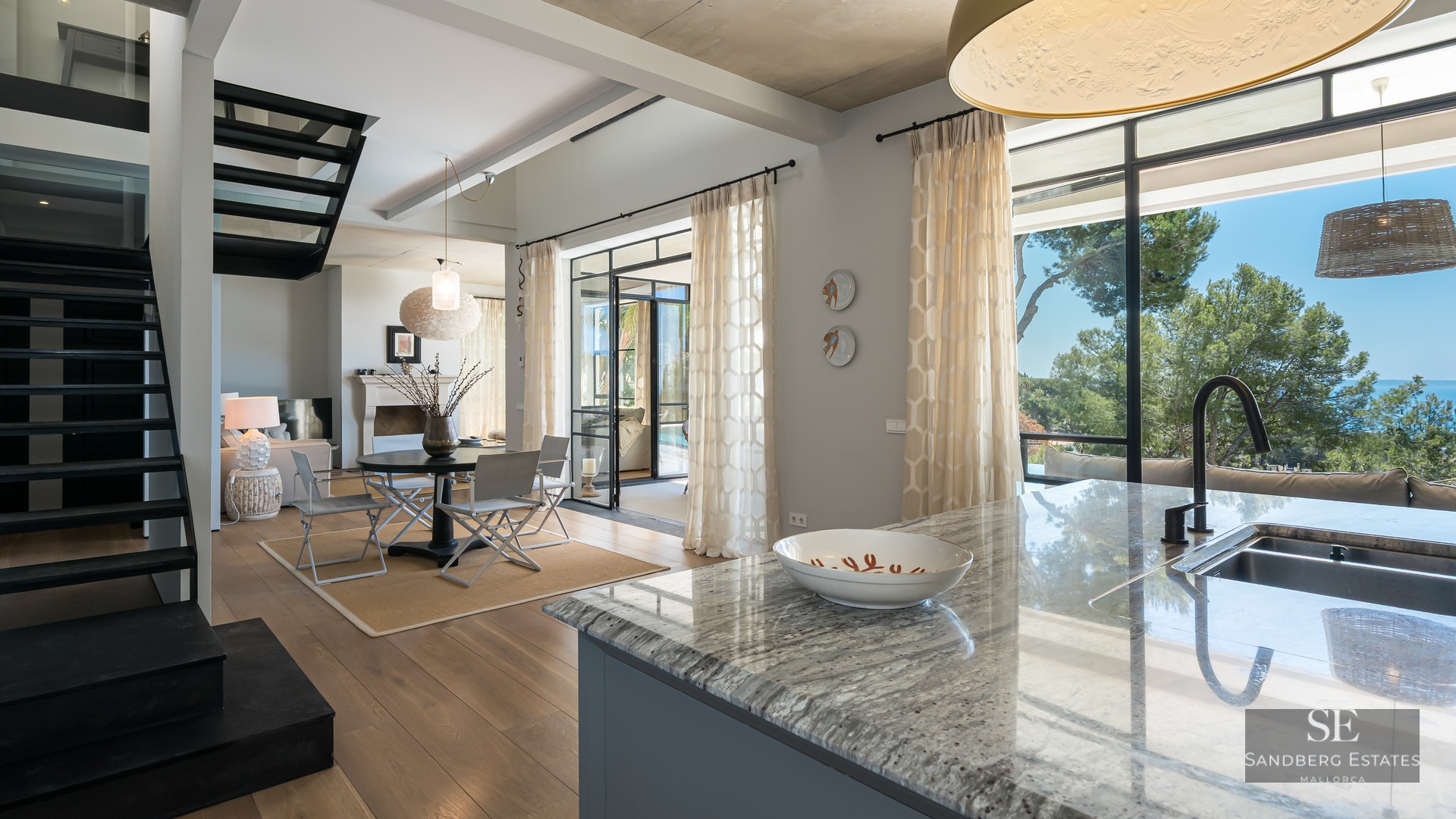 Designer living area featuring a marble island, black metal staircase, and large windows overlooking the sea.