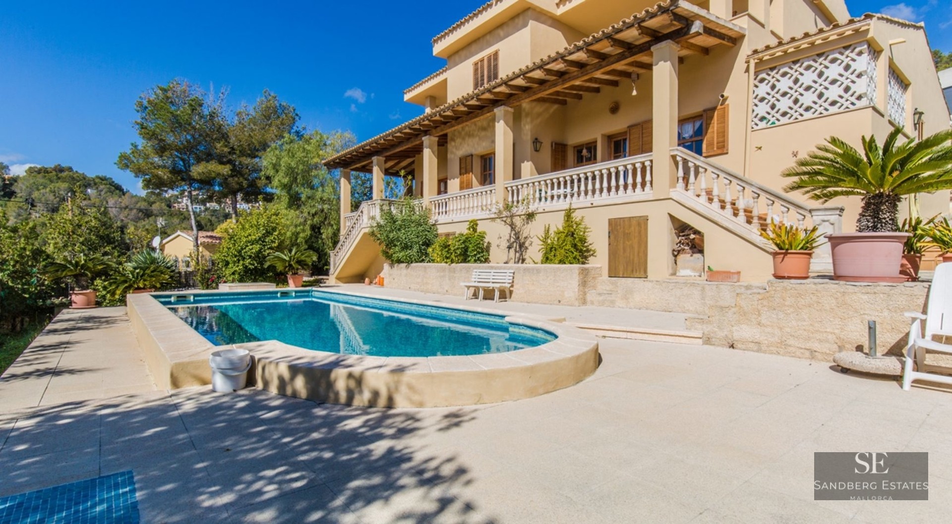 Large swimming pool in front of a multi-story Mediterranean villa with a terrace and lush green hills under a blue sky.