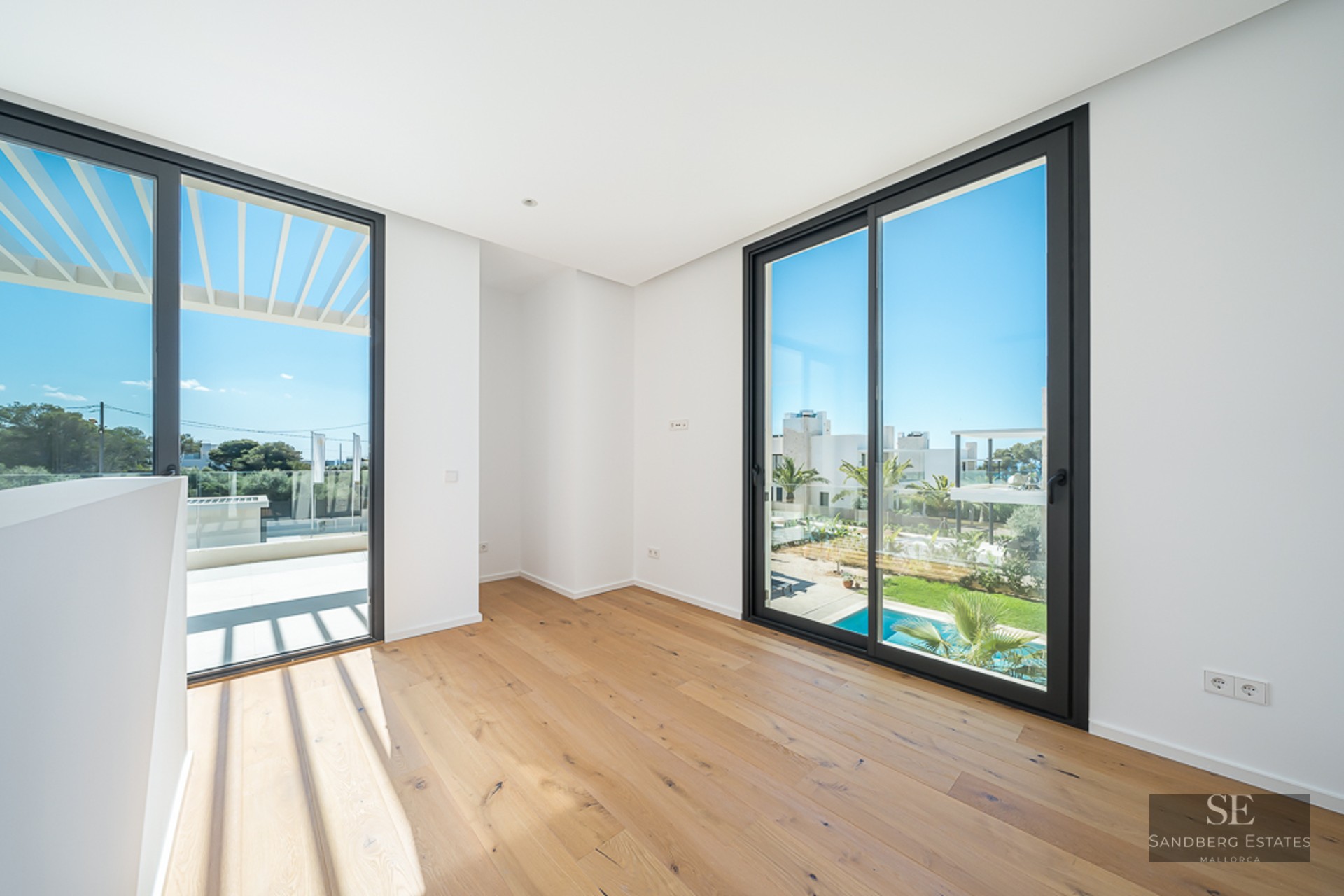 Empty room with light oak floors, white walls, and large glass sliding doors overlooking a terrace and swimming pool.