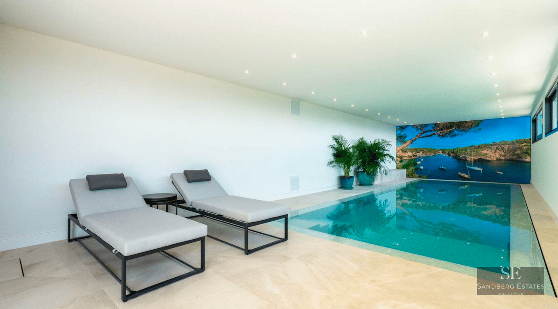 Modern indoor swimming pool with turquoise water, grey sun loungers, and a large scenic coastal wall mural.