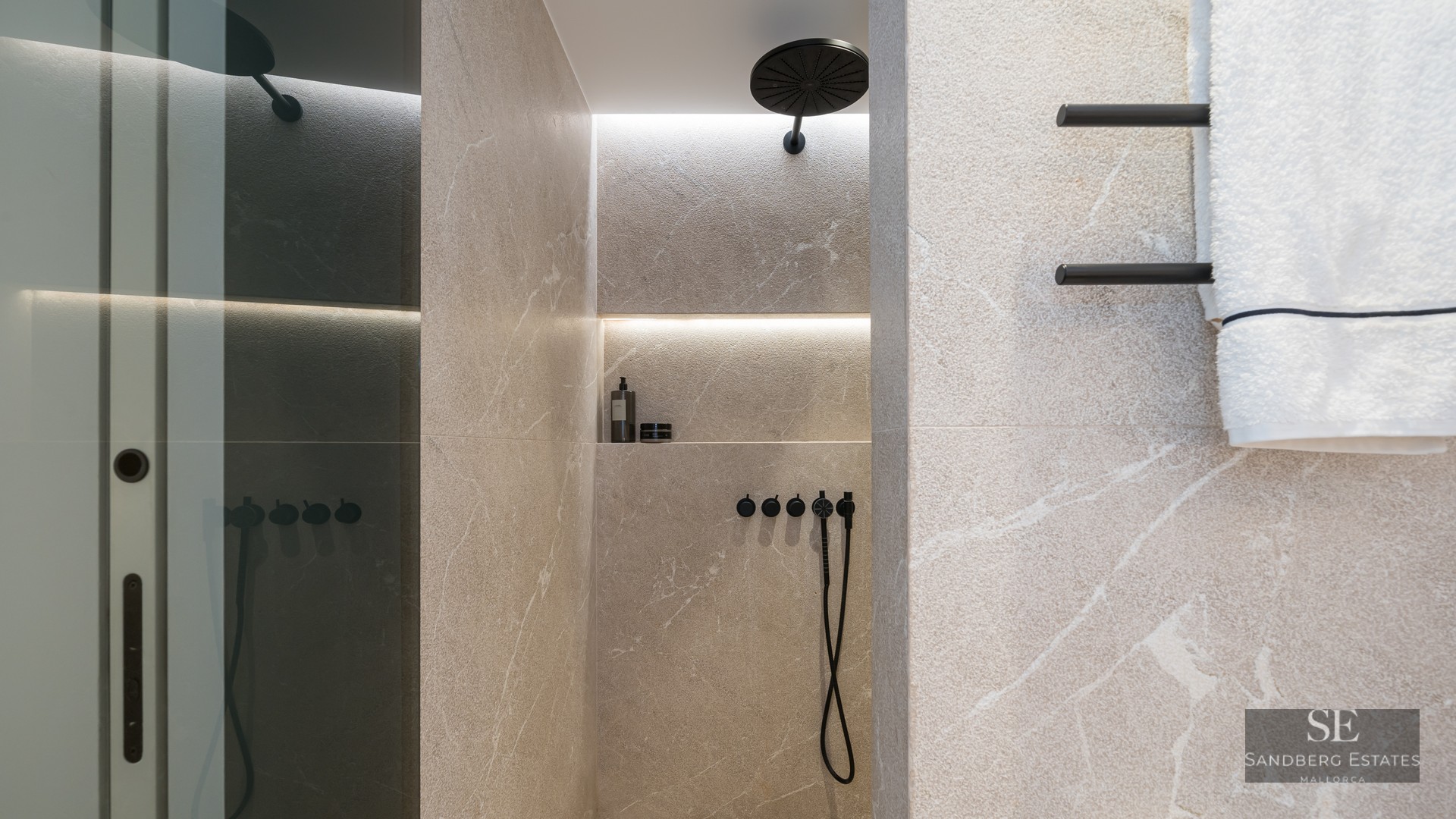 Walk-in shower with beige stone tiles, a matte black rain shower head, and an illuminated recessed niche on veined stone.