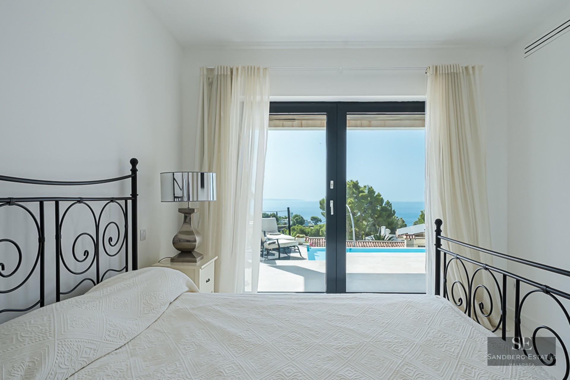 White bedroom with a black wrought iron bed and glass doors opening to a sunny terrace with a pool and sea view.
