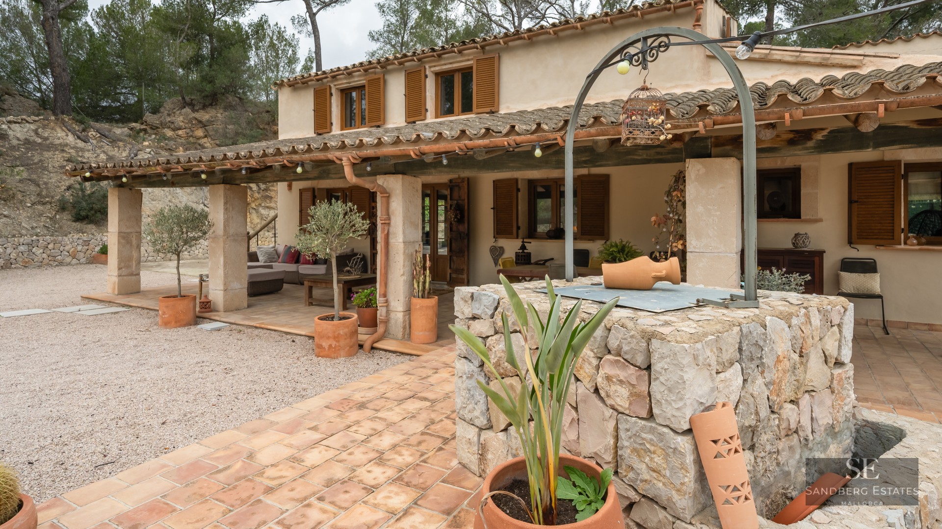 Facade of a rustic house with a covered porch, wooden shutters, and a stone well in the foreground.