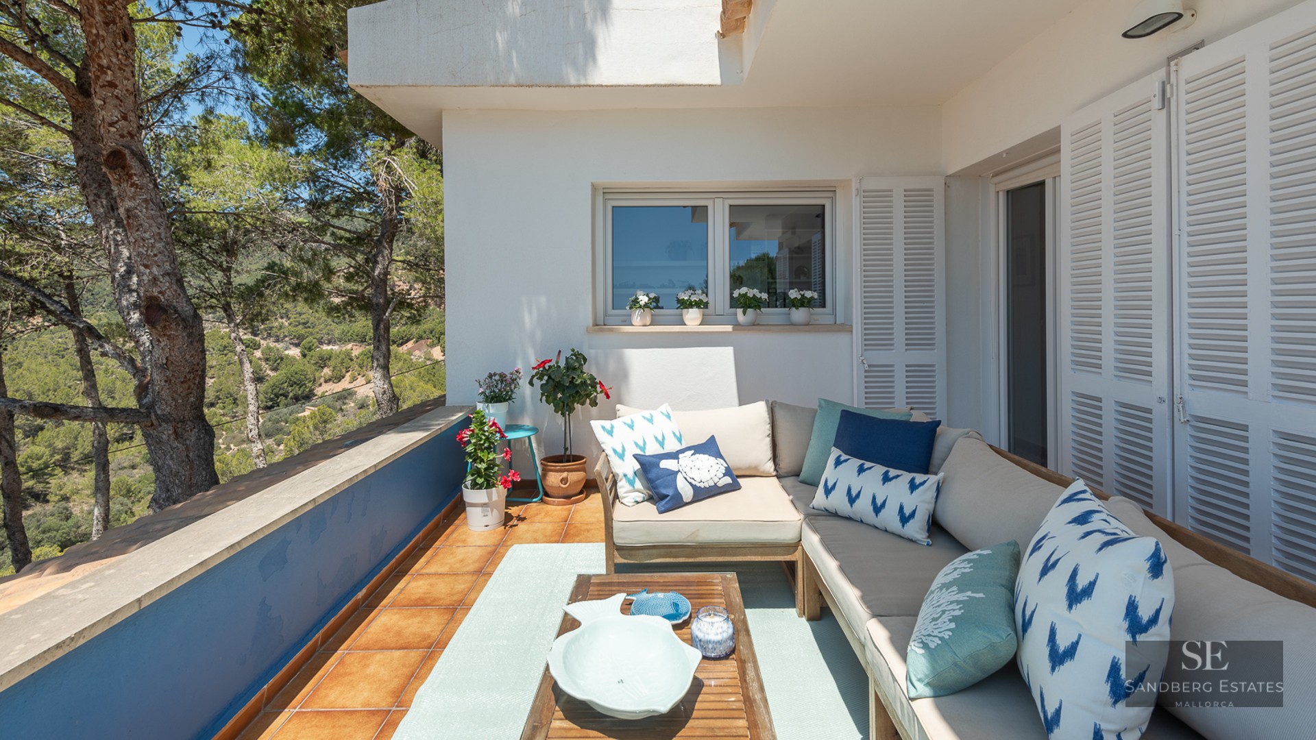 Outdoor terrace with a large L-shaped sofa, blue cushions, and a wooden table overlooking a pine forest valley.