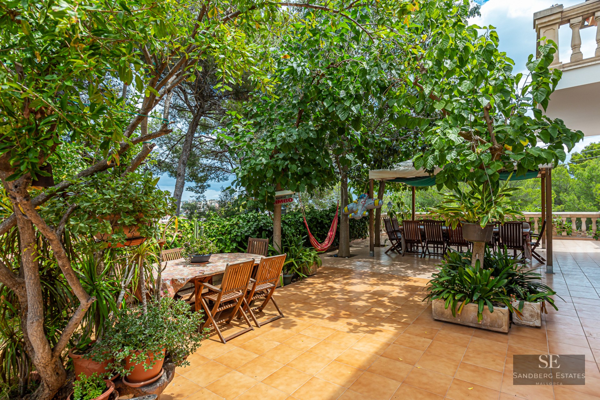 Large tiled terrace surrounded by mature trees featuring two wooden dining areas and a red hammock in the shade.