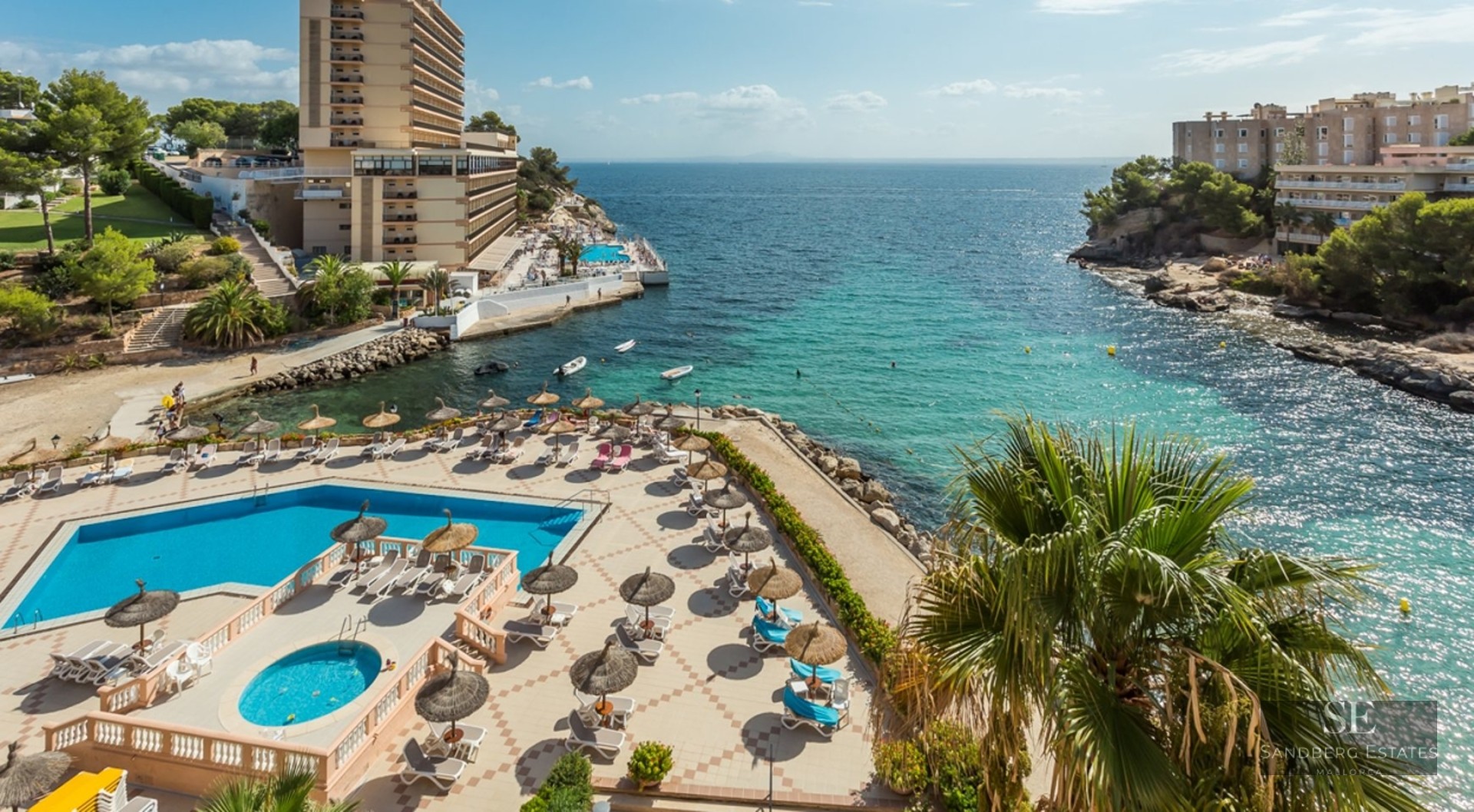 Aerial view of a swimming pool area with sunbeds and umbrellas overlooking a clear turquoise bay and rocky coastline.