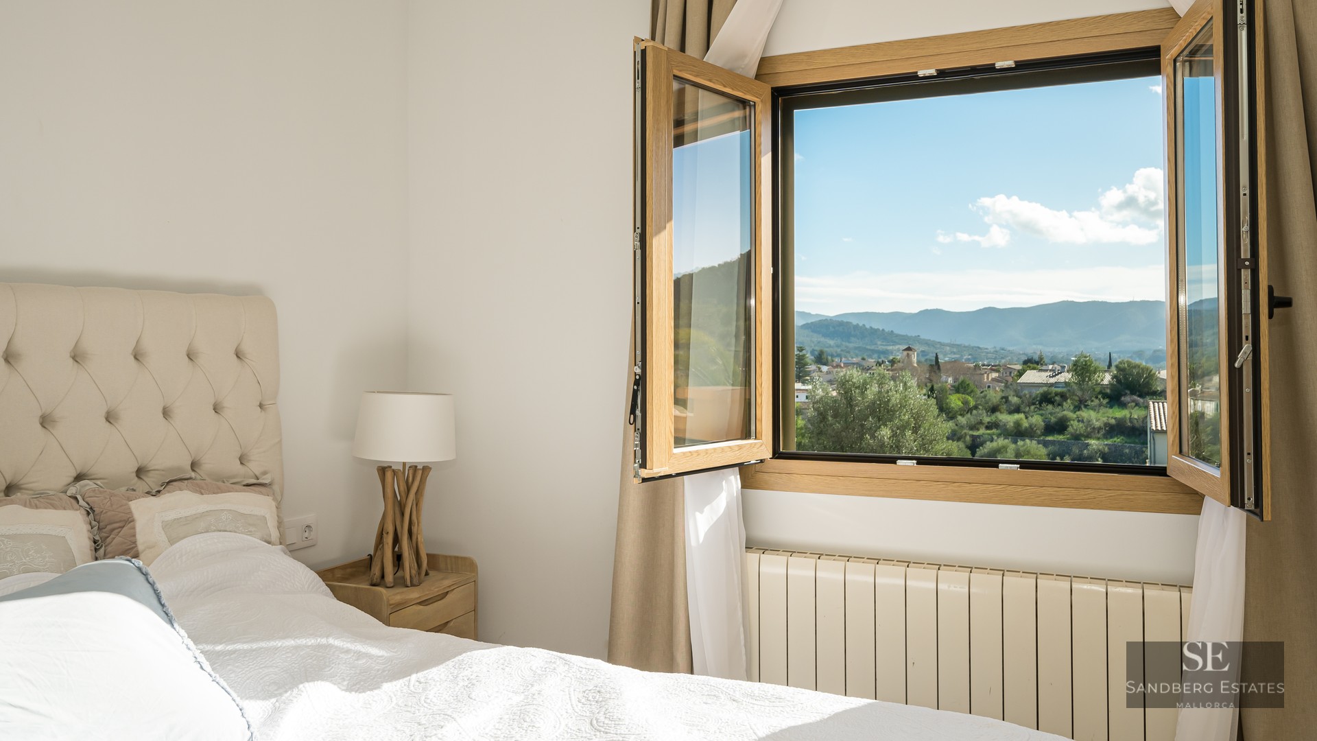Cozy bedroom with tufted headboard and open window showing panoramic views of a village and mountains.