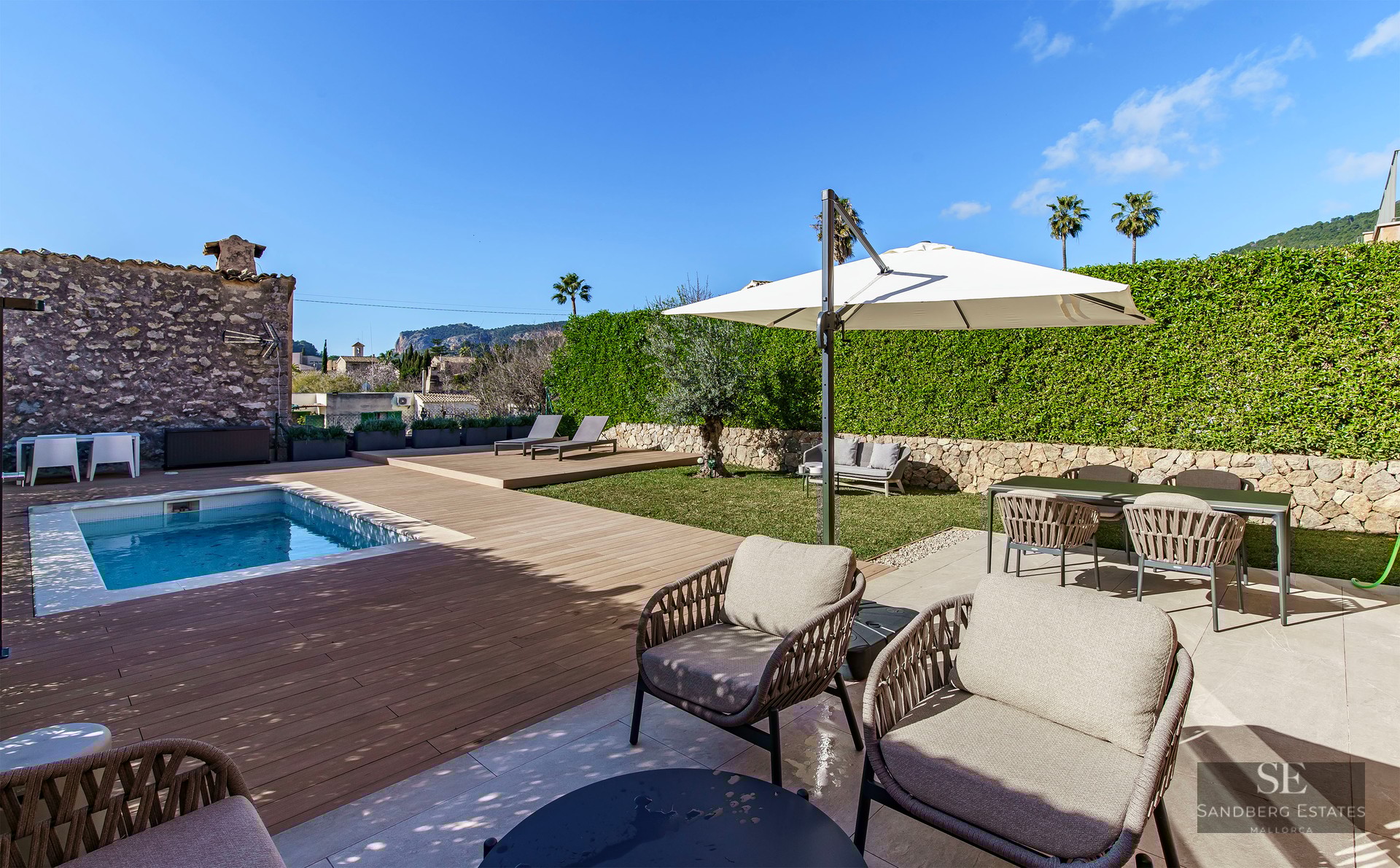 Modern swimming pool with wooden deck, stylish outdoor furniture, and lush green hedges under a clear blue sky.