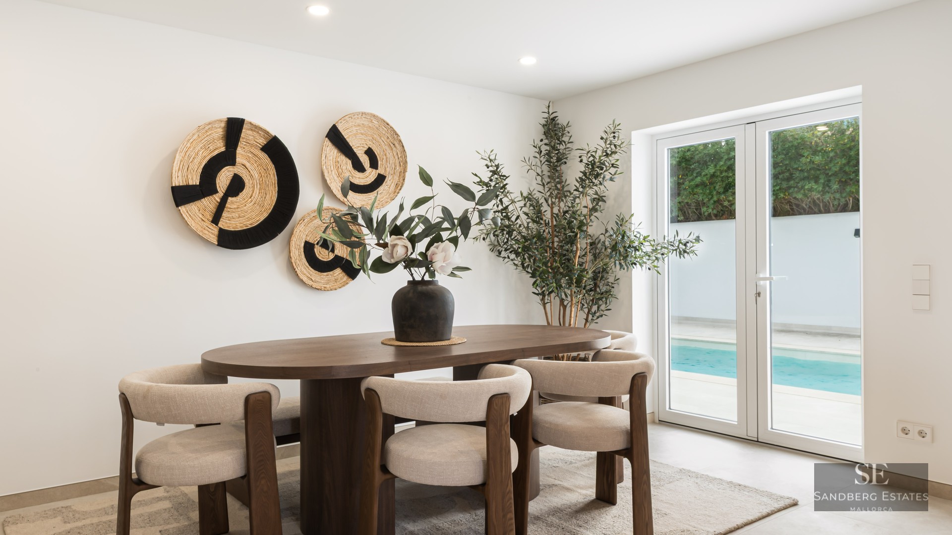 Oval wooden dining table with beige chairs in front of glass doors showing a swimming pool.