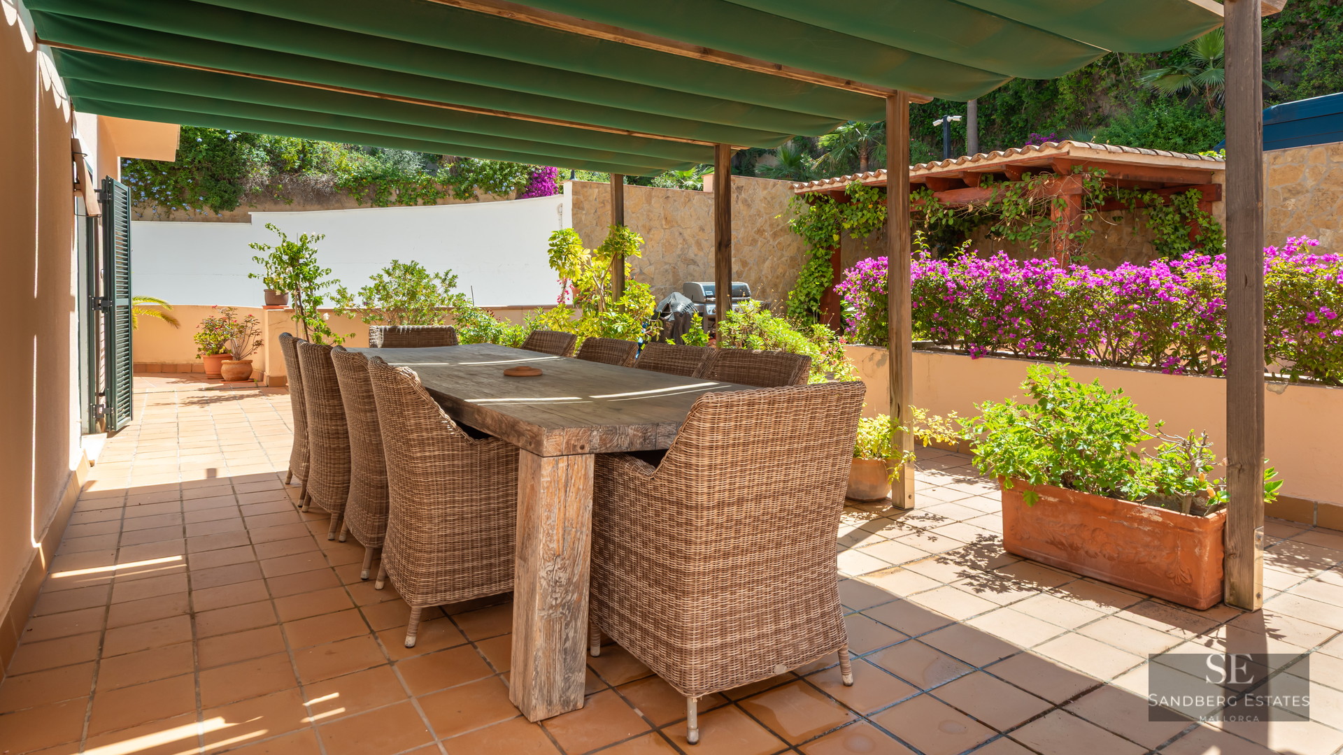Large wooden dining table with wicker chairs on a sunny terracotta terrace under a green awning.