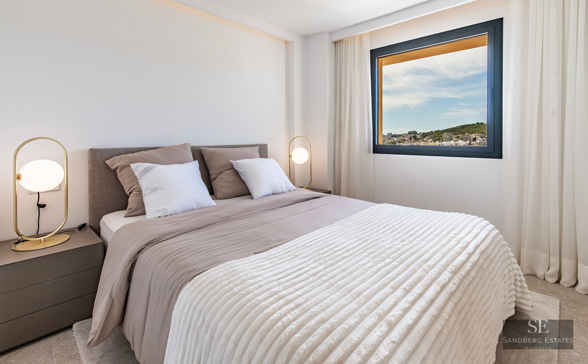 Modern bedroom with a large bed, gold designer lamps, and a window overlooking a city landscape.