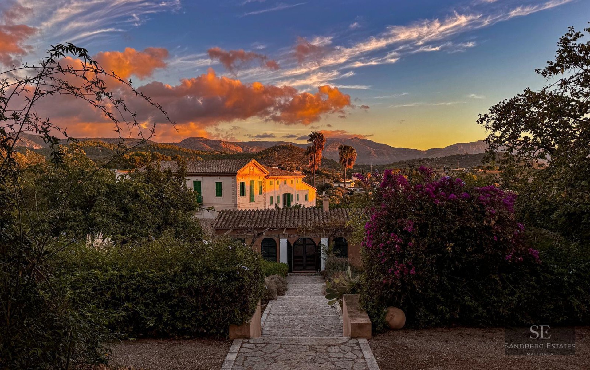 Scenic view of a white Mediterranean villa with terracotta roof at sunset, surrounded by lush gardens and mountains.