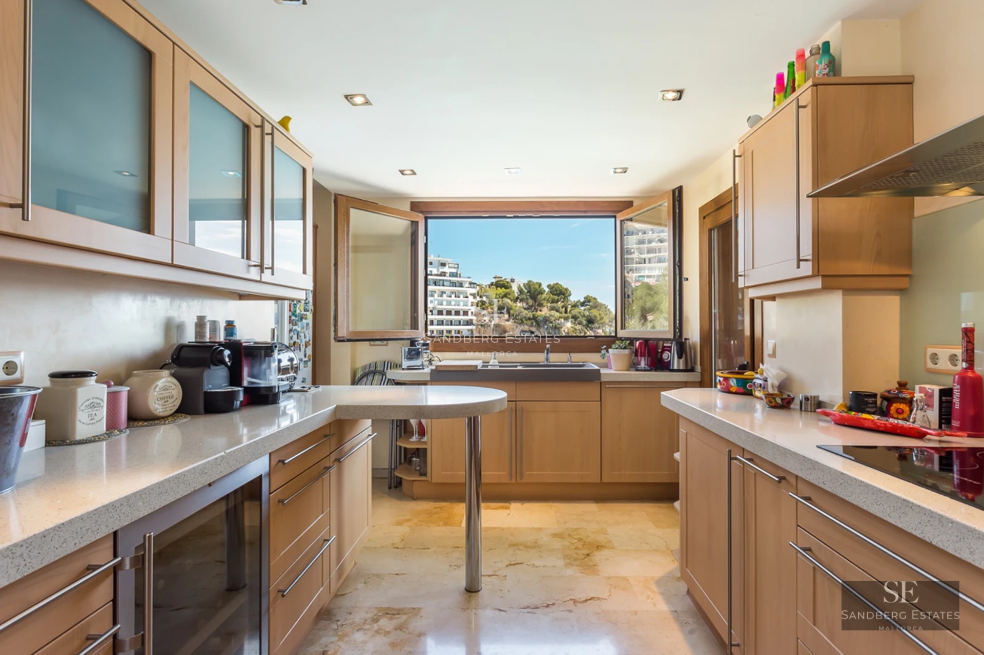 Modern kitchen with light wood cabinets, marble floors, and a large window overlooking a coastal landscape.