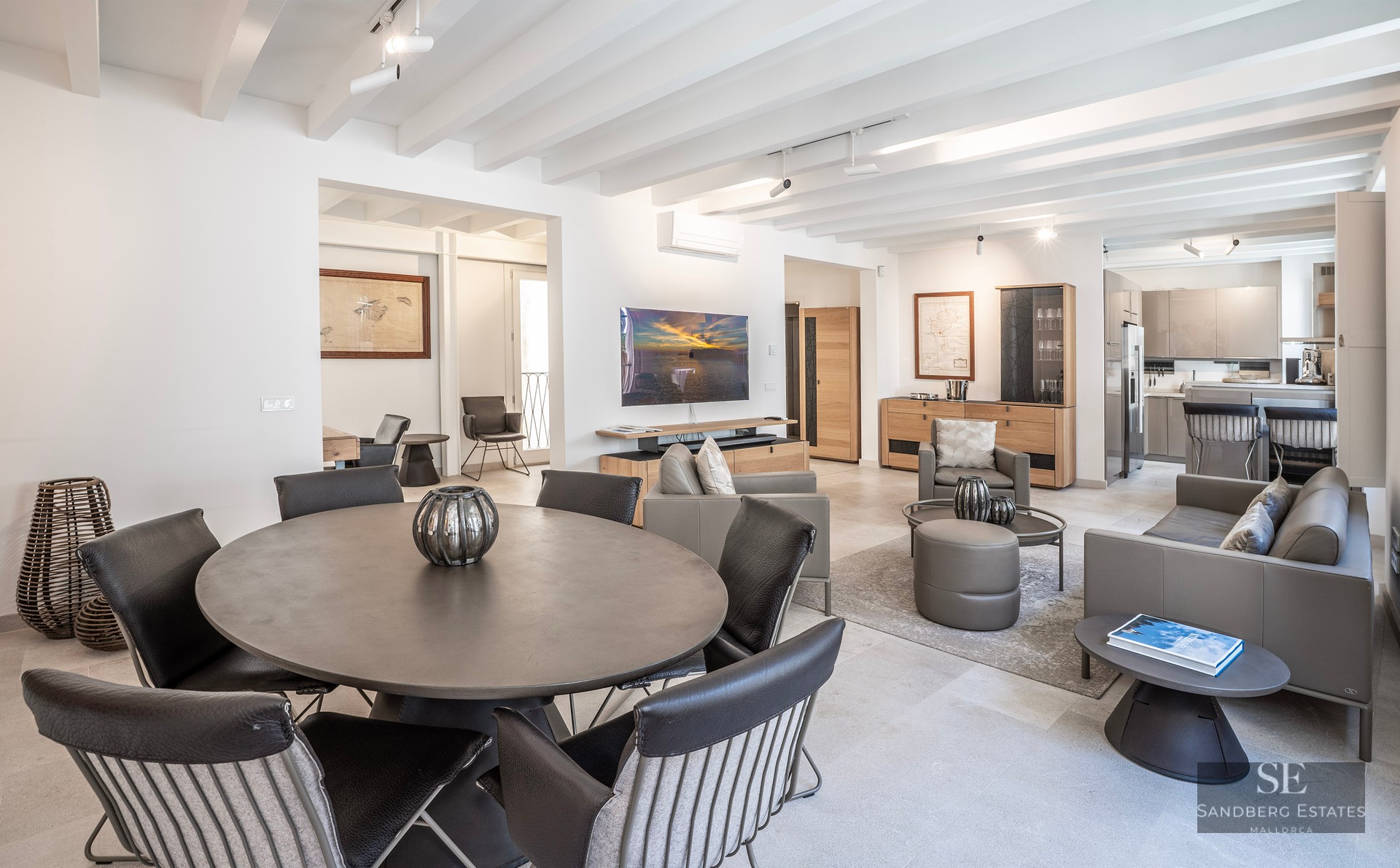 Salon moderne avec table ronde, canapés en cuir, plafond à poutres blanches et télévision murale.