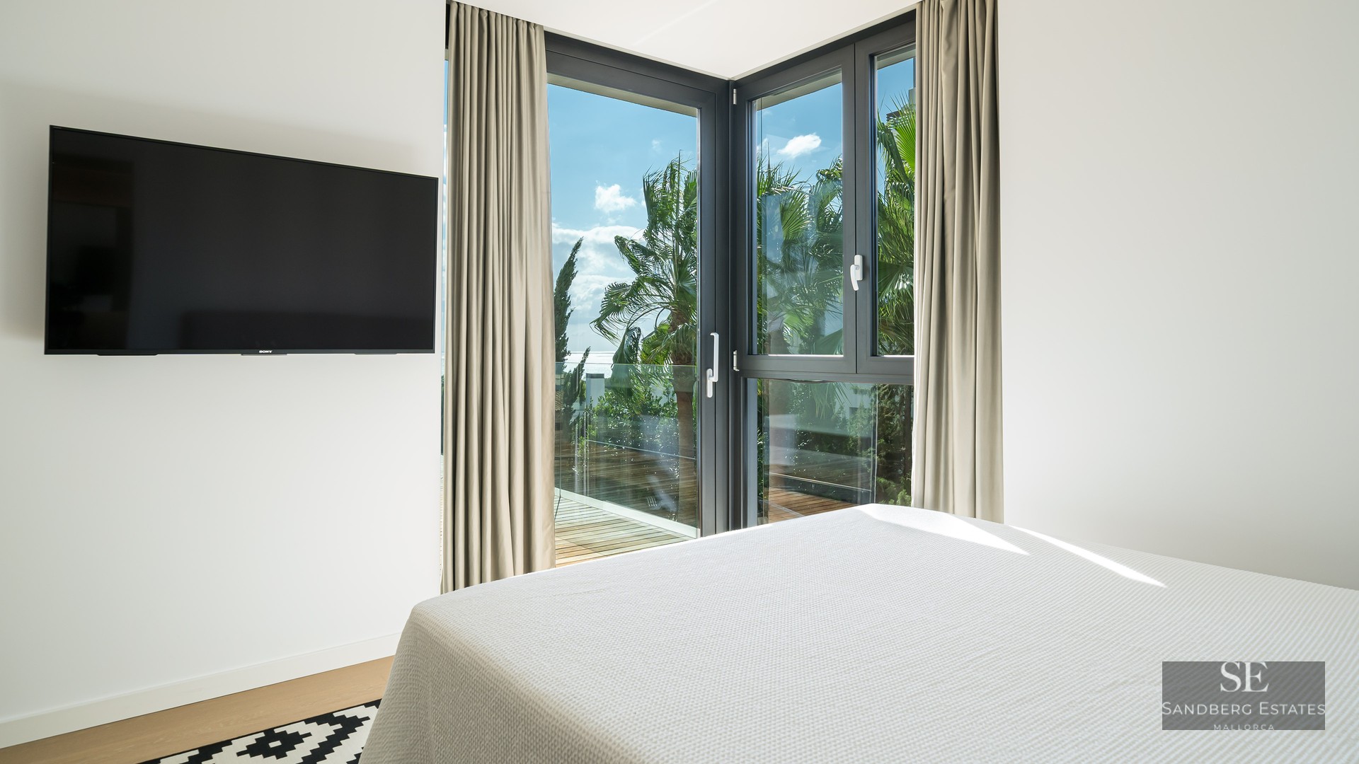Bright bedroom with a large bed, wall-mounted TV, and corner windows looking out to palm trees and a sunny sky.