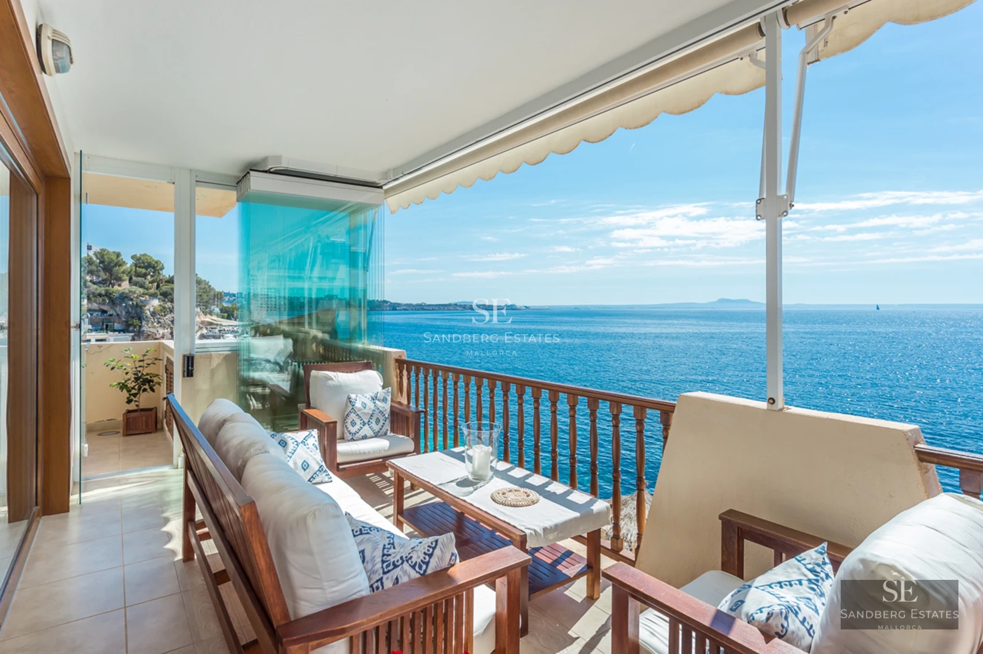 A sunlit balcony with wooden furniture and white cushions overlooking a sparkling blue sea under a clear sky.