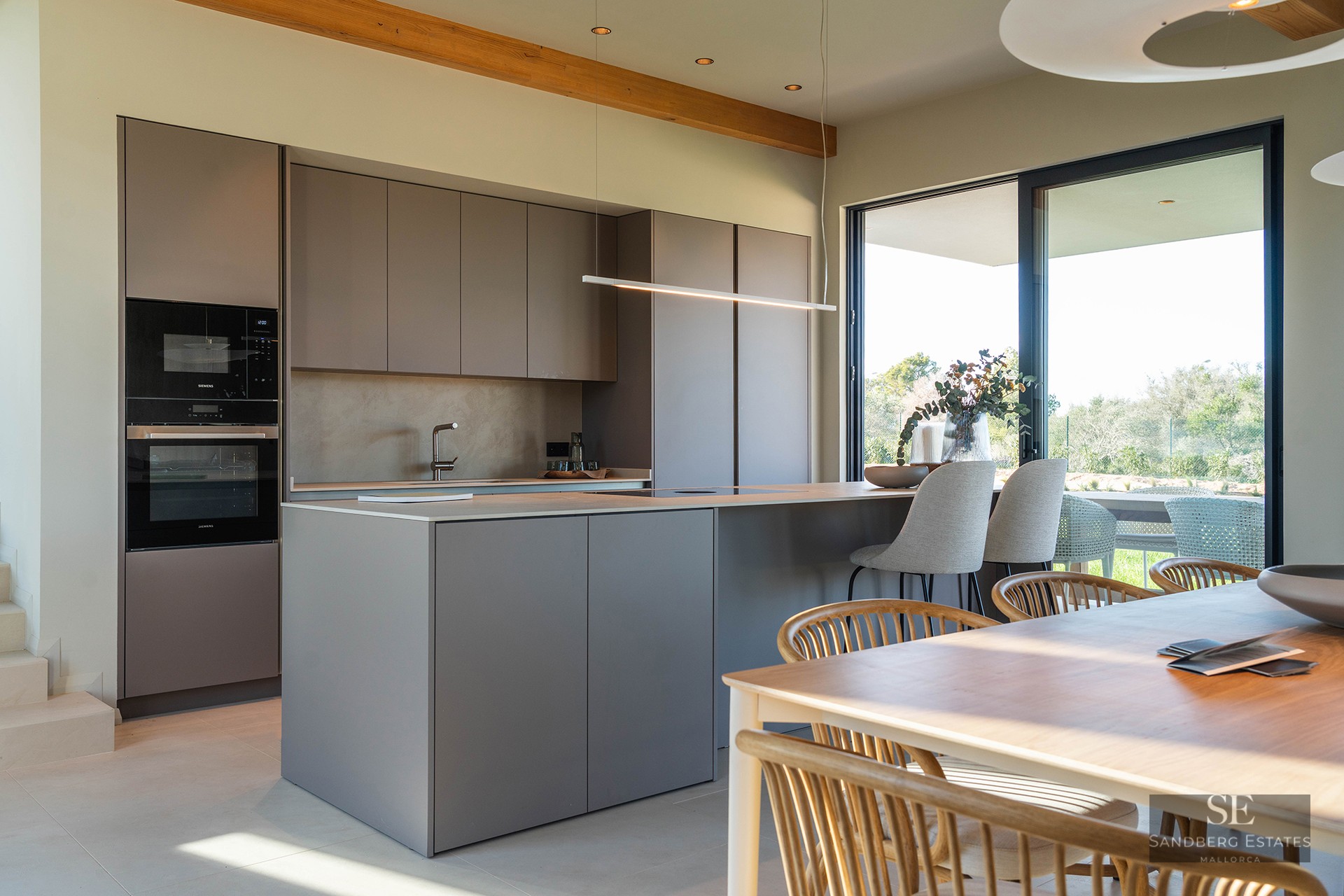 Minimalist kitchen with taupe island, integrated appliances, and wooden dining table under exposed beams.