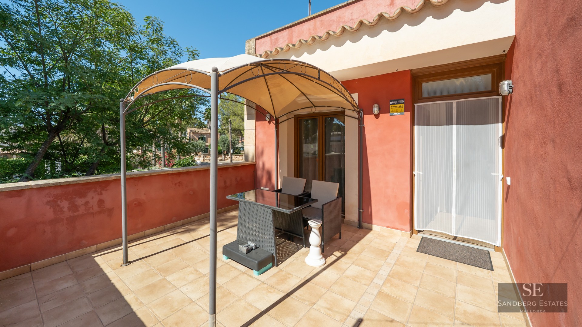 Sunny terrace with terracotta walls, a tiled floor, and a metal gazebo over a table and two chairs.