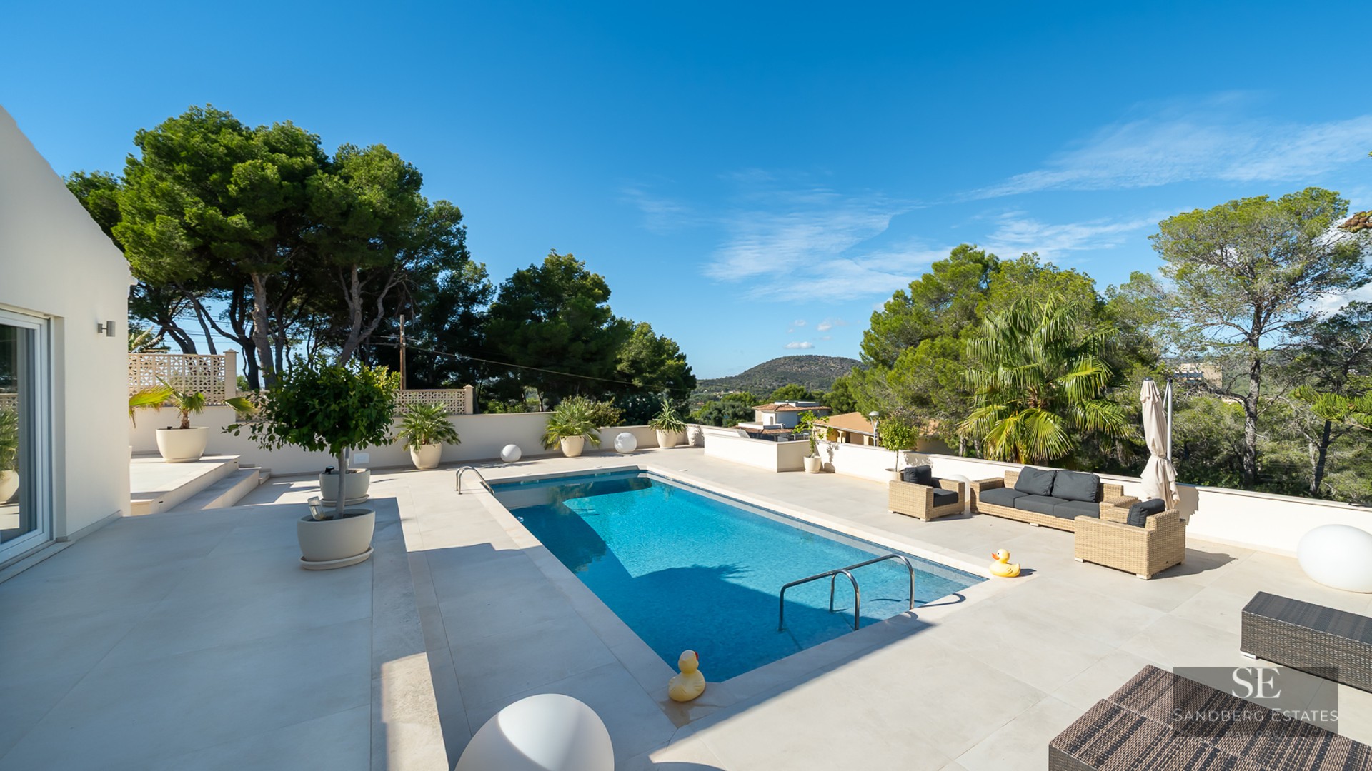 Piscine bleue rectangulaire sur terrasse blanche avec mobilier de jardin et vue sur la forêt méditerranéenne.