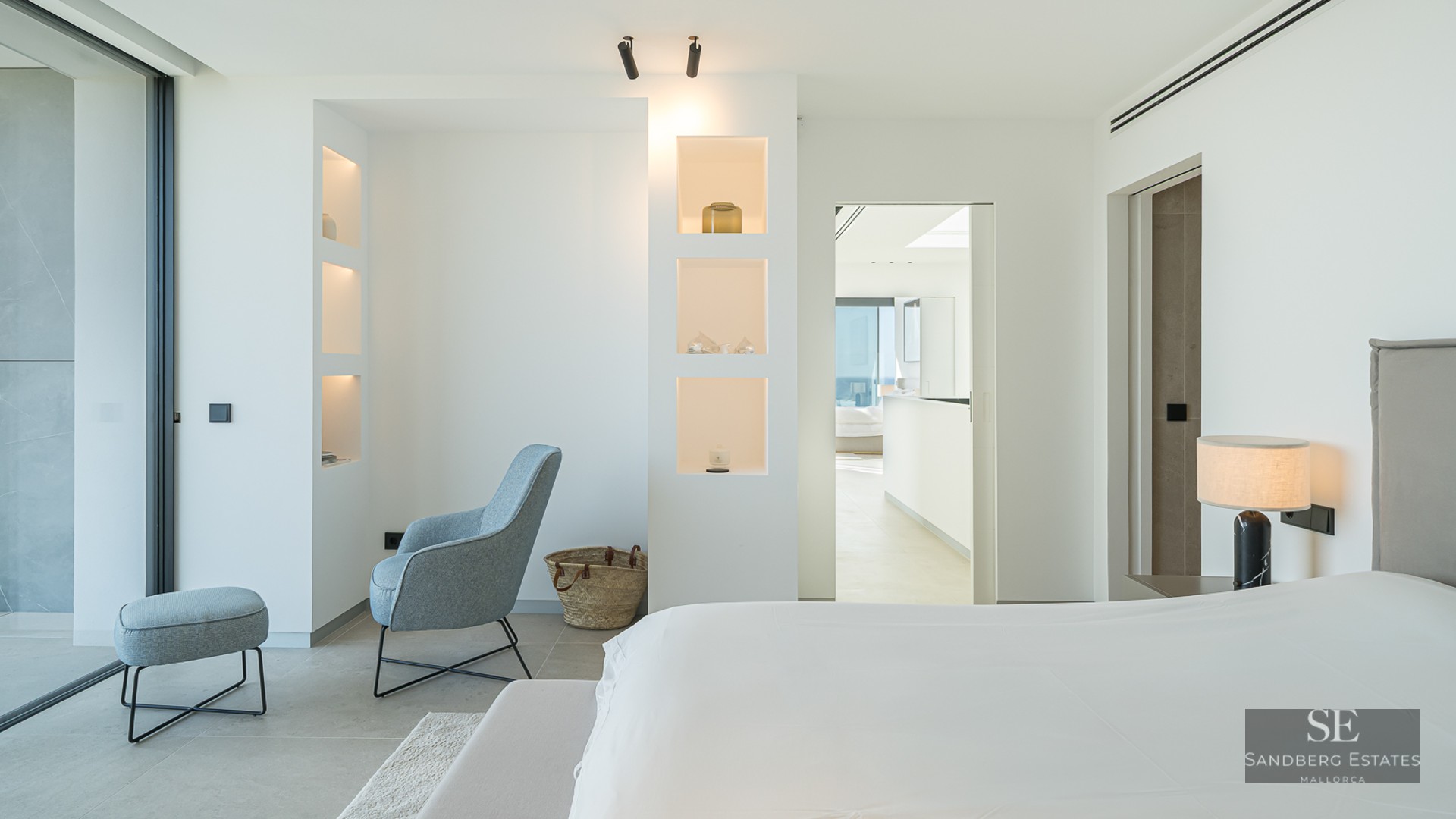 Bright modern master bedroom featuring a grey bed, blue designer armchair, and built-in illuminated wall shelving.