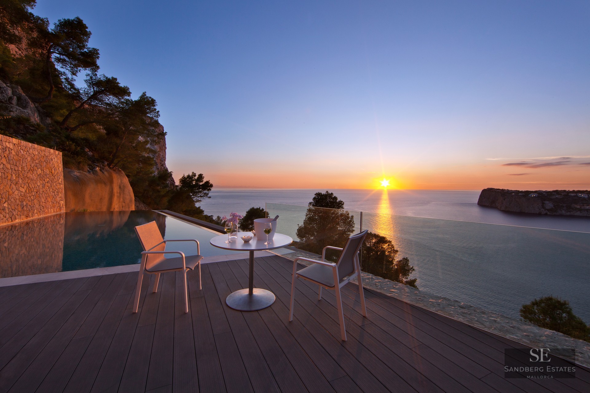 Sunset view from a luxury terrace with an infinity pool, wooden decking, and white modern outdoor seating.
