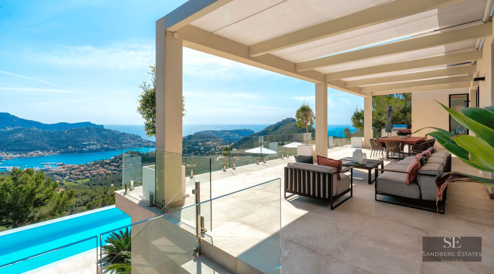 Large modern terrace with grey sofas, glass railings, and a stunning view of a blue bay and green hills.