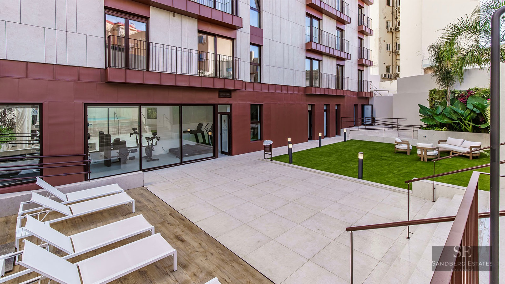 Courtyard with stone tiles, sun loungers, a lawn area with sofas, and a glass-walled gym at a modern building.