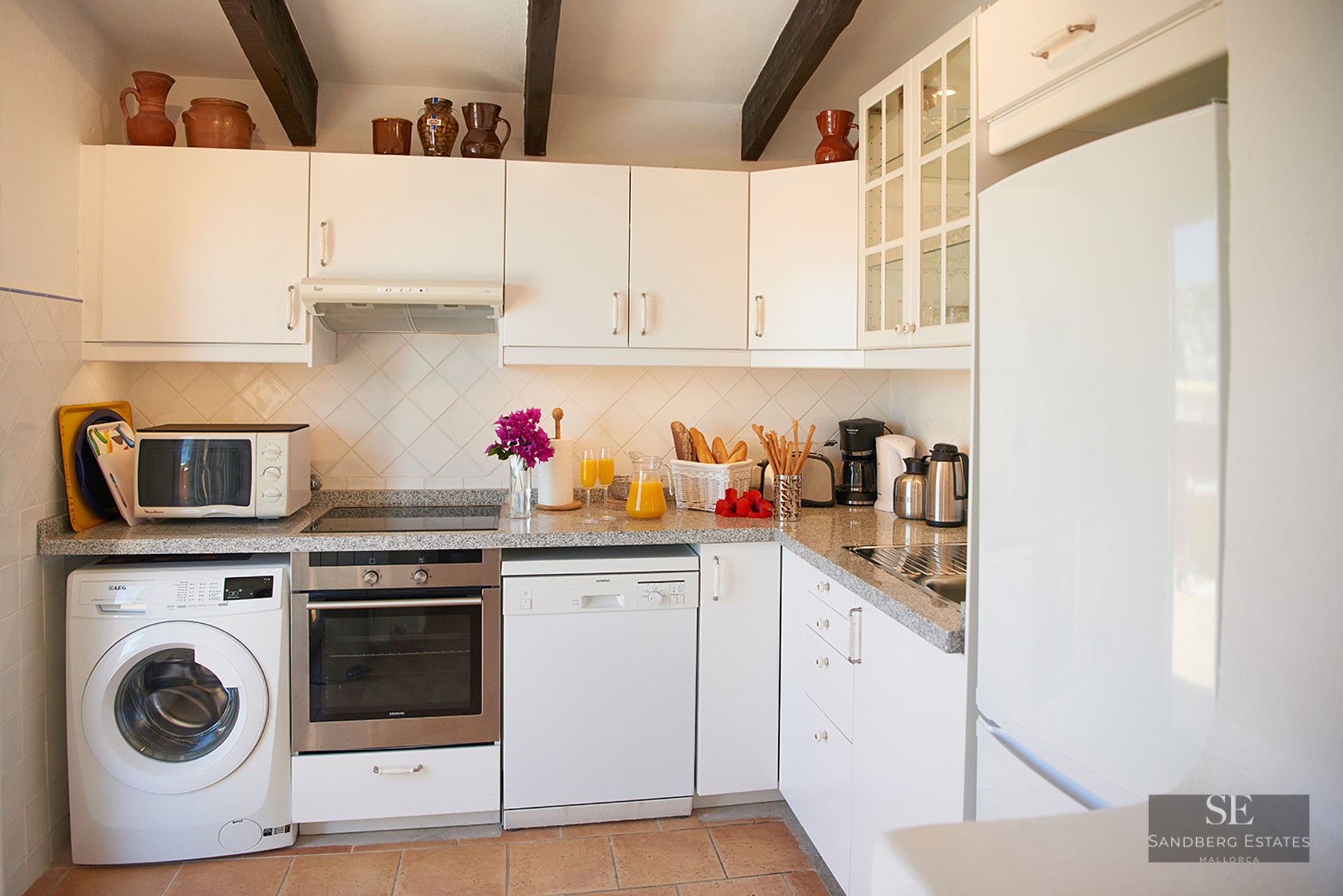 Bright kitchen with white cabinets, dark wood beams, terracotta floor, and modern appliances.