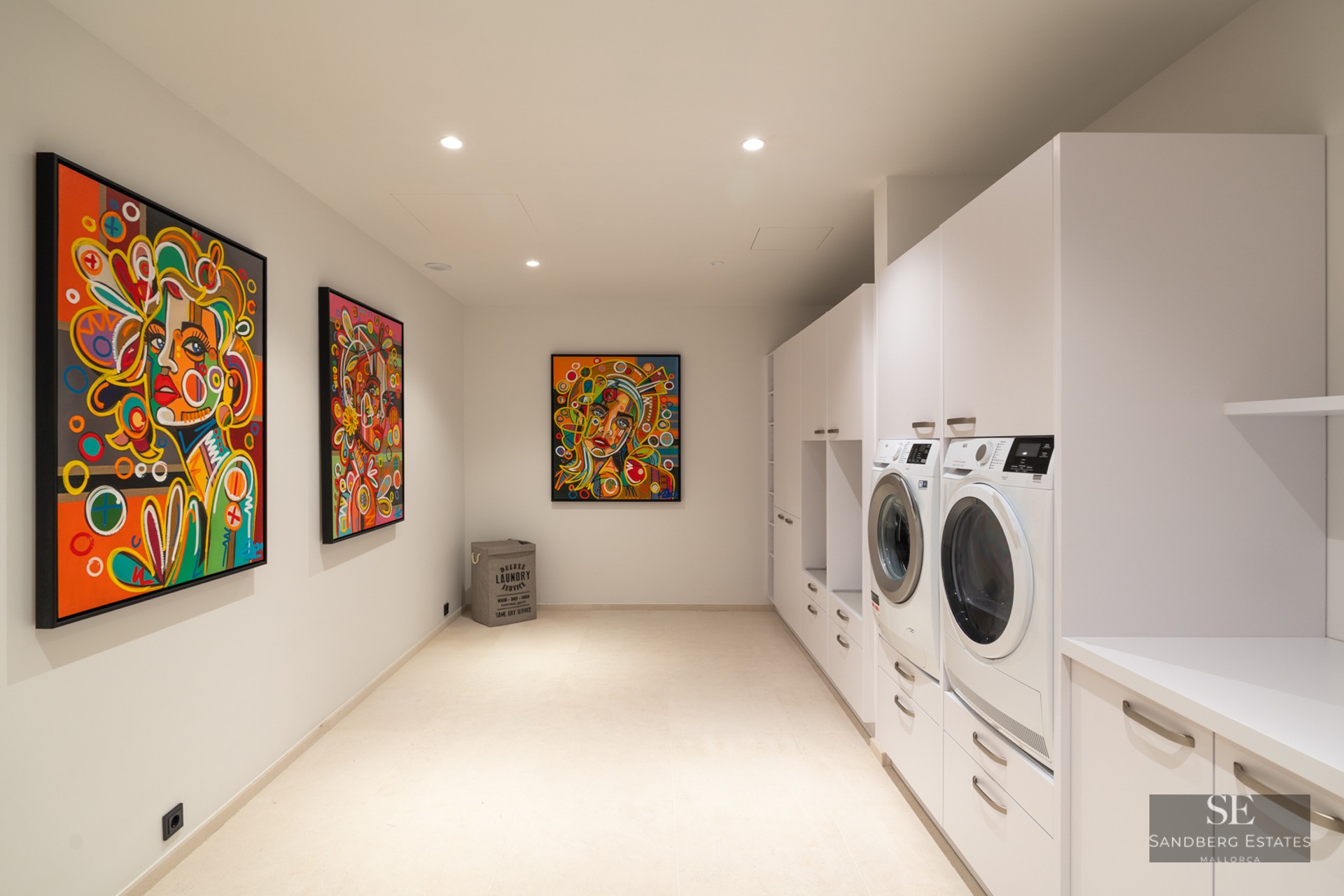 Modern white laundry room with washer, dryer, minimalist cabinetry, and three vibrant colorful pop-art portraits.