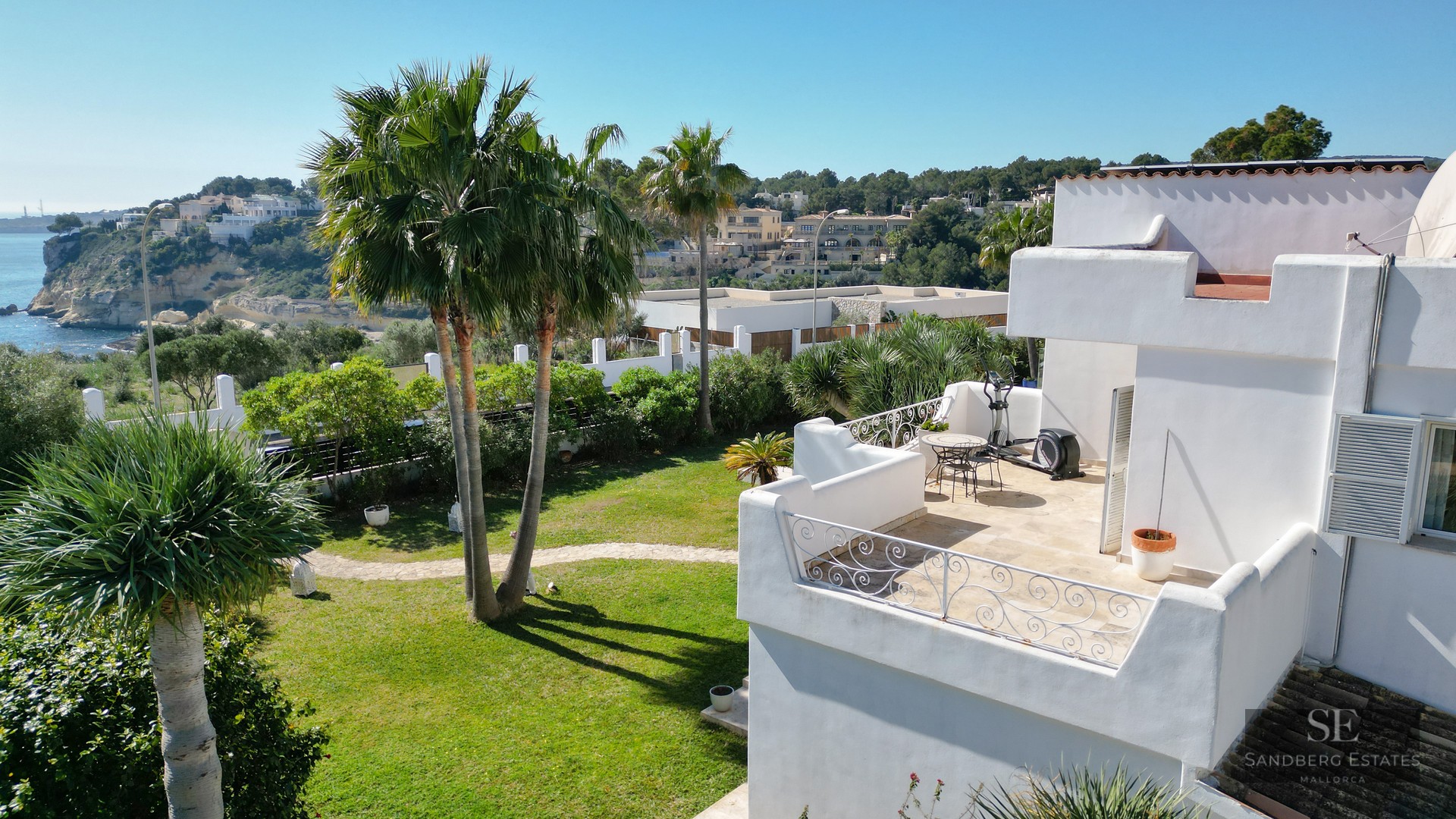 High-angle view of a white villa with a terrace, garden with palm trees, and views of the blue sea and cliffs in the distance.