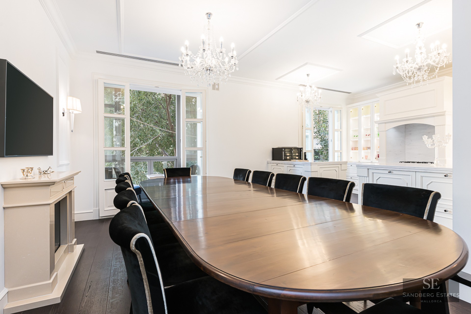 Large wooden dining table with black velvet chairs under crystal chandeliers in a bright, white room with a fireplace.