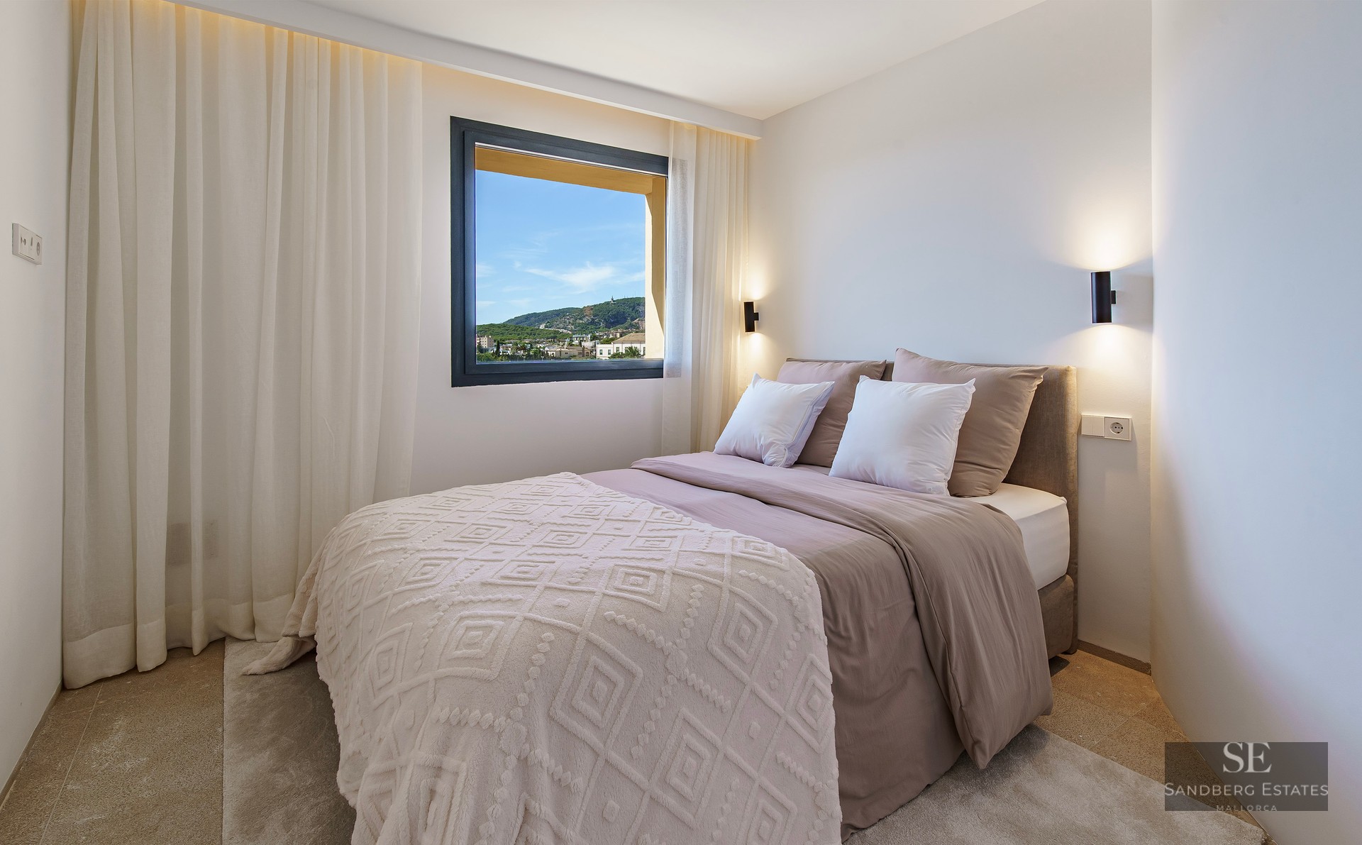 Modern bedroom featuring a double bed, white patterned throw, and a window framing a view of green hills and blue sky.