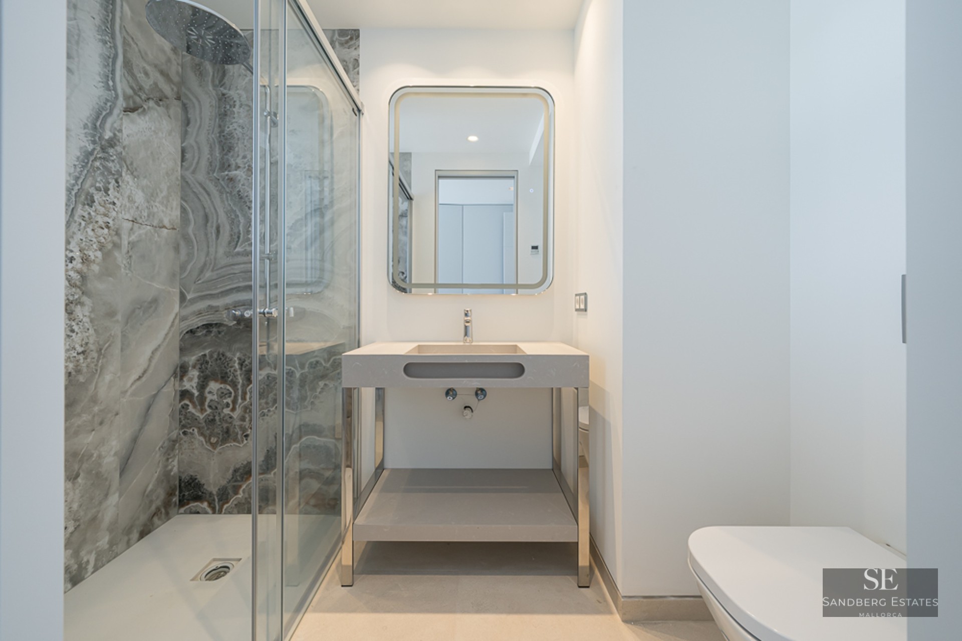Modern bathroom with a glass walk-in shower featuring marble-patterned walls, a backlit mirror, and a minimalist sink.