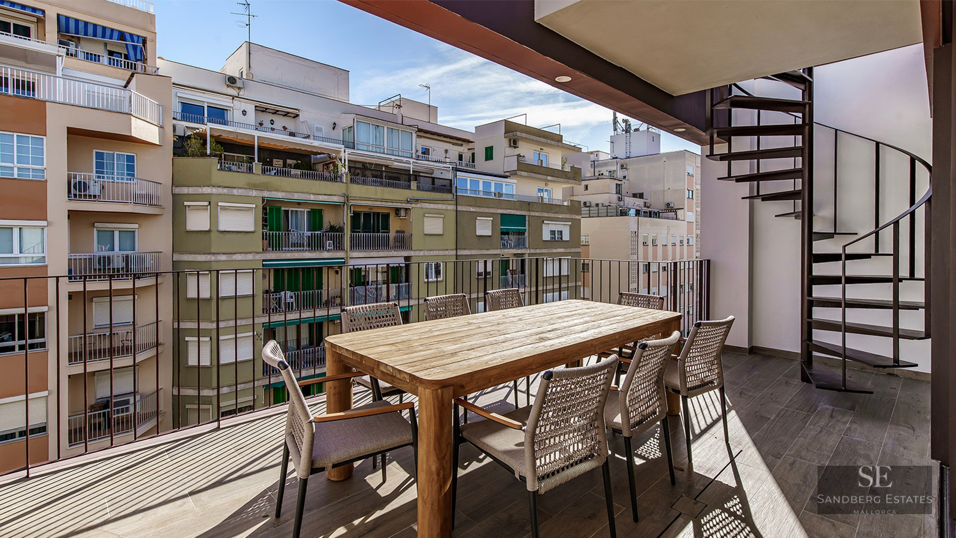 Sunlit terrace with wooden dining table, woven chairs, and black spiral staircase against a city backdrop.