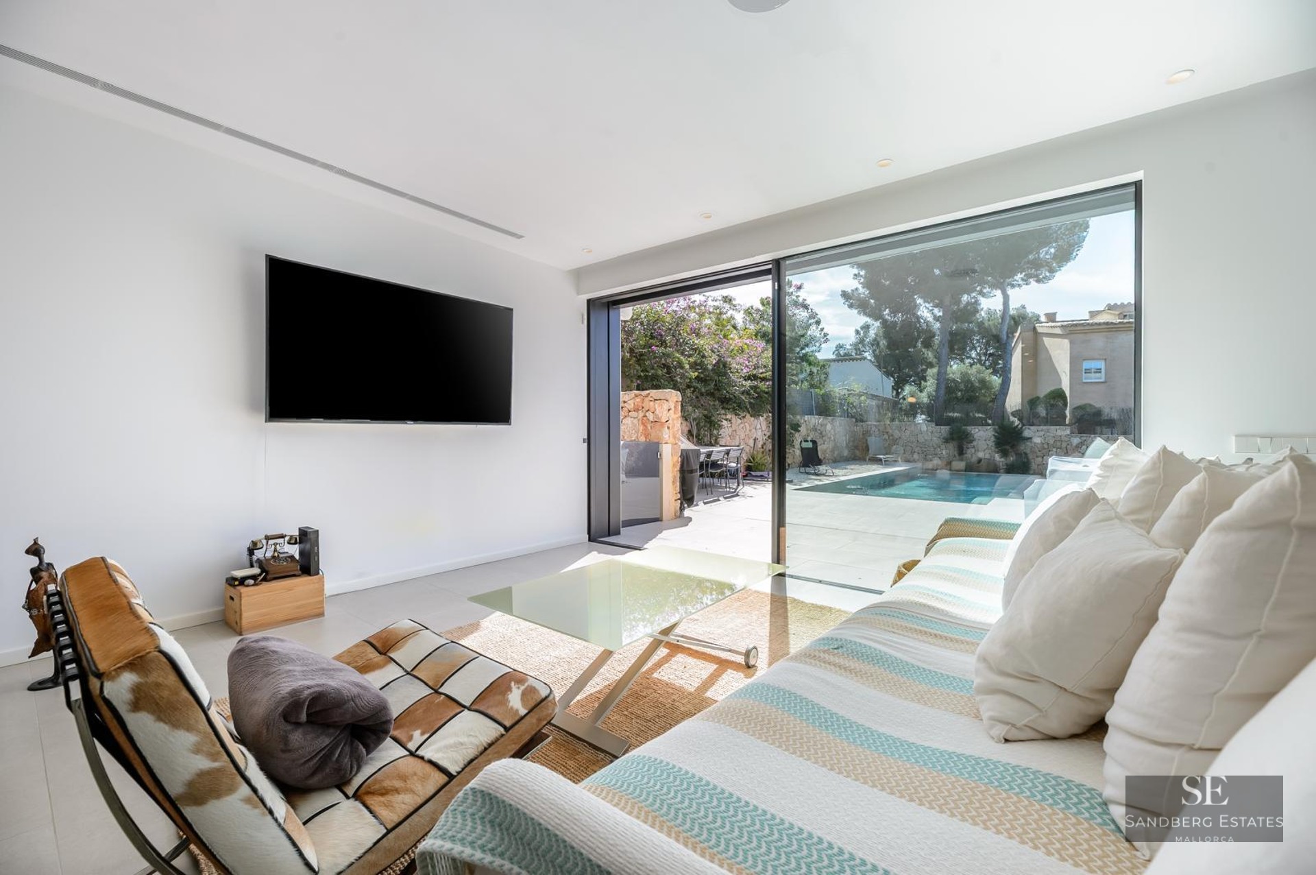 Bright living room with a striped sofa, large TV, and floor-to-ceiling glass doors leading to a pool terrace.