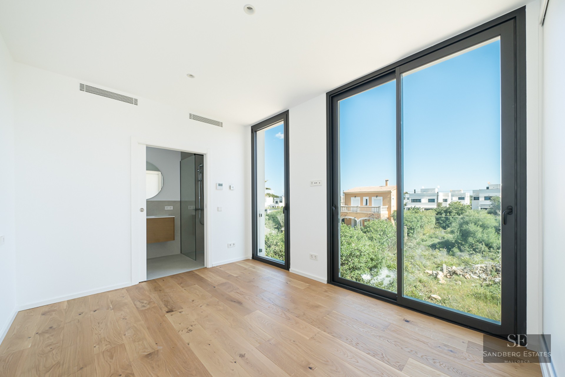 Empty modern bedroom with light wood floors, large black-framed windows overlooking greenery, and an open ensuite bathroom.