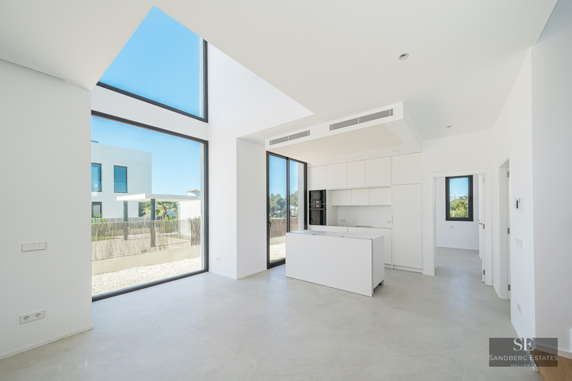 Minimalist white kitchen with an island, double-height floor-to-ceiling windows, and bright natural light.