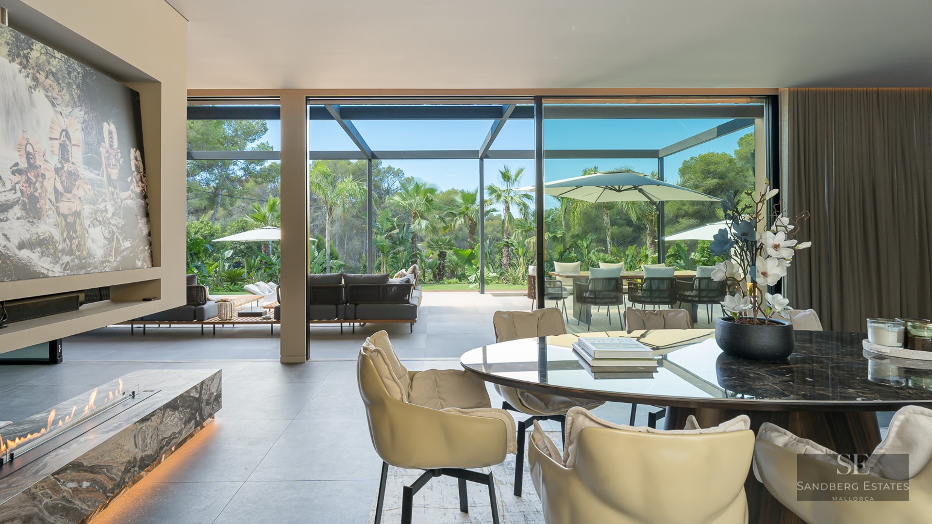 Modern dining room with a marble table and linear fireplace, opening through glass doors to a lush tropical terrace.