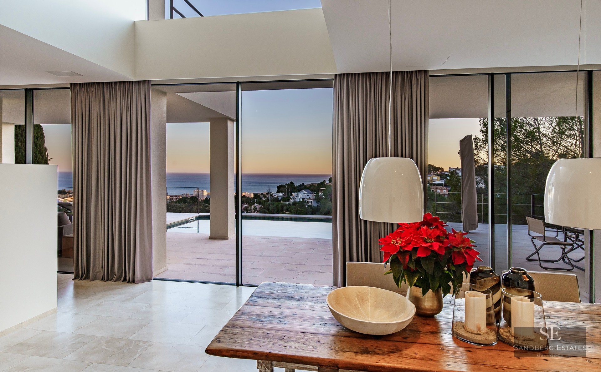 Interior dining area with a rustic wooden table overlooking an outdoor terrace with an infinity pool and sea view.