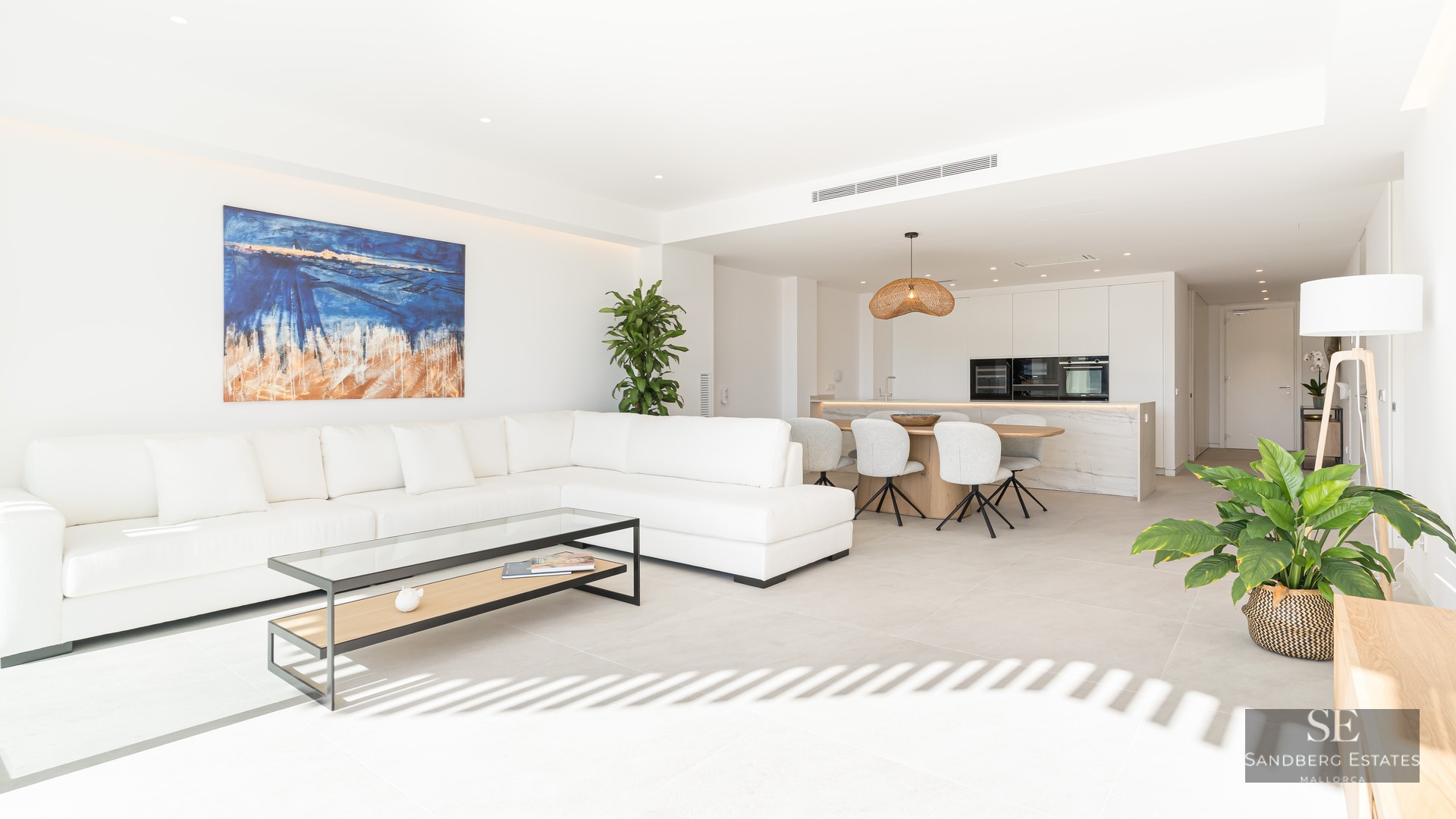 Bright living room with white L-shaped sofa, glass coffee table, abstract art, and integrated kitchen and dining space.
