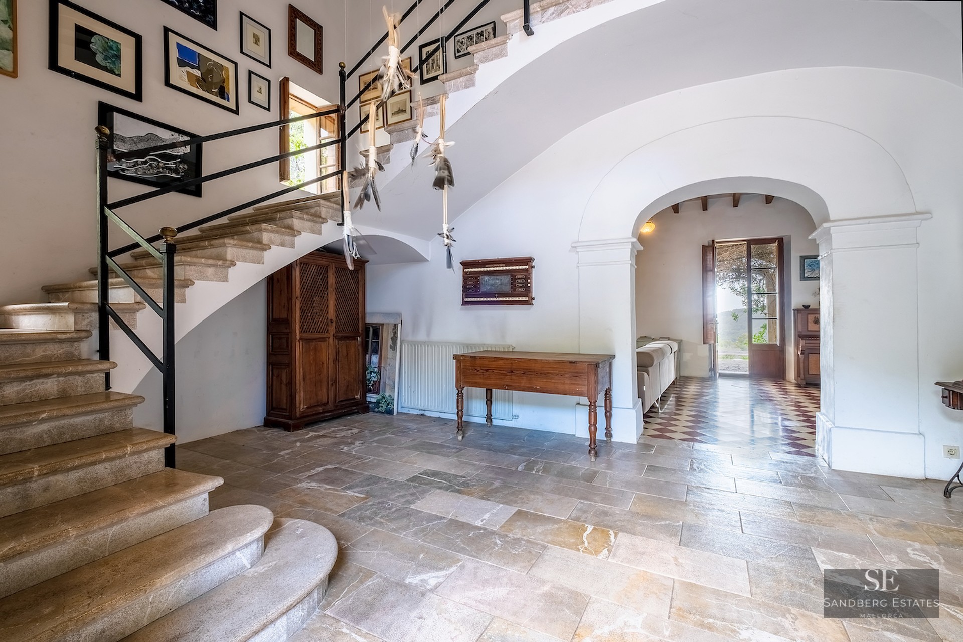 A grand entrance hall with a stone staircase, antique wooden furniture, and a gallery wall under an arched doorway.
