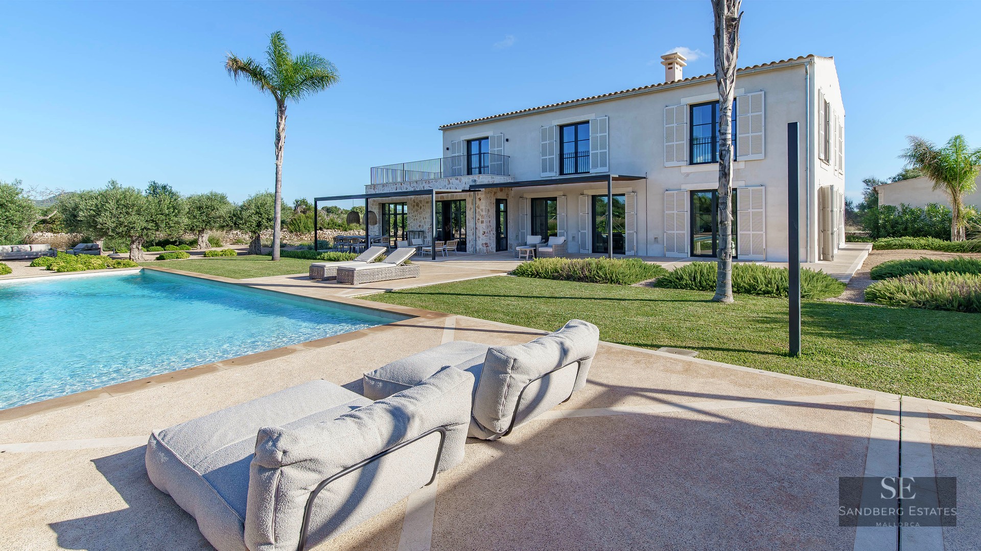 Large turquoise swimming pool in front of a stone villa with palm trees and green lawn under a clear blue sky.