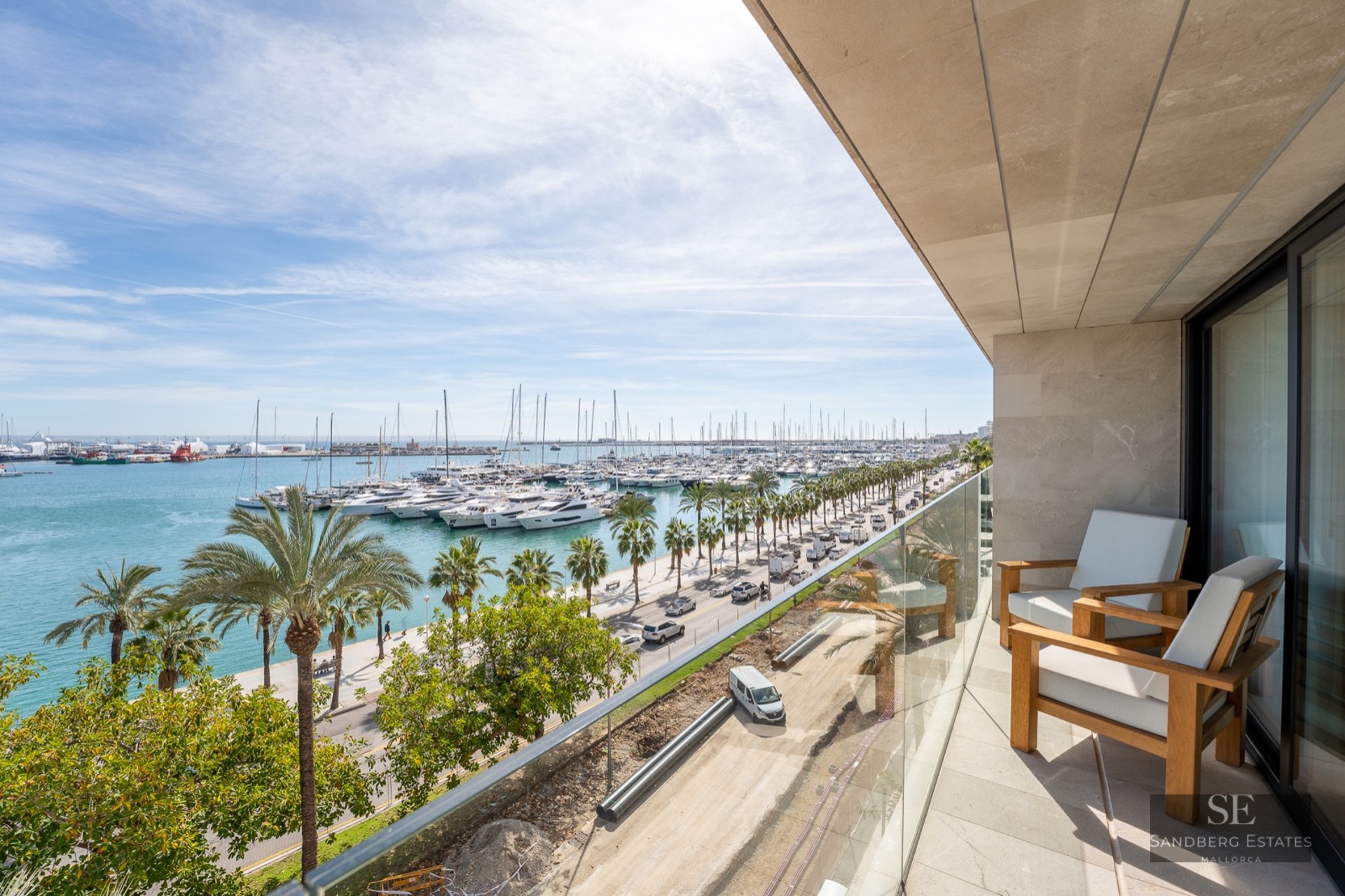 Modern balcony with wooden armchairs and glass railing overlooking a marina filled with yachts and palm trees.
