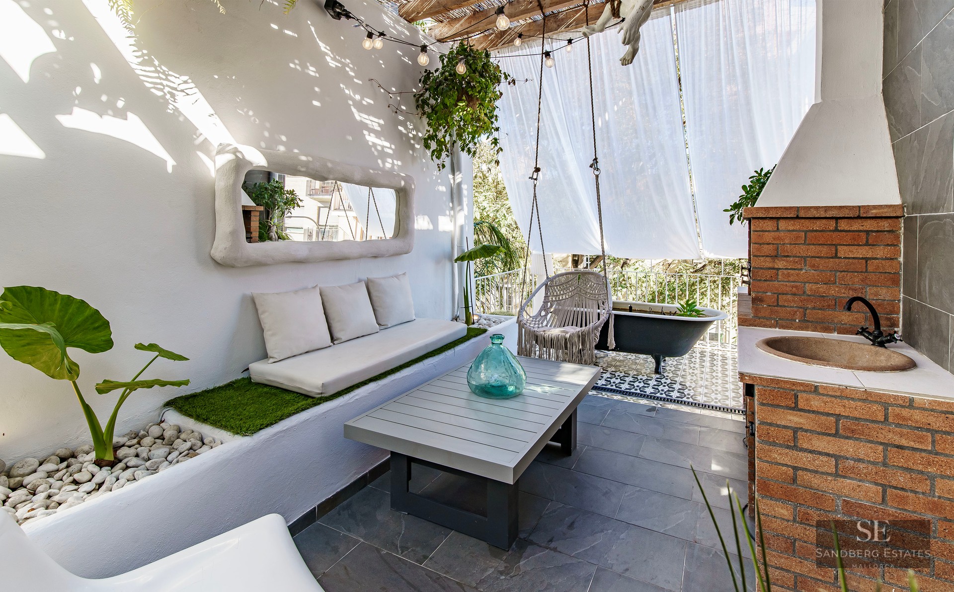 Sun-drenched white terrace with built-in sofa, macrame swing, brick outdoor sink, and a black clawfoot bathtub.